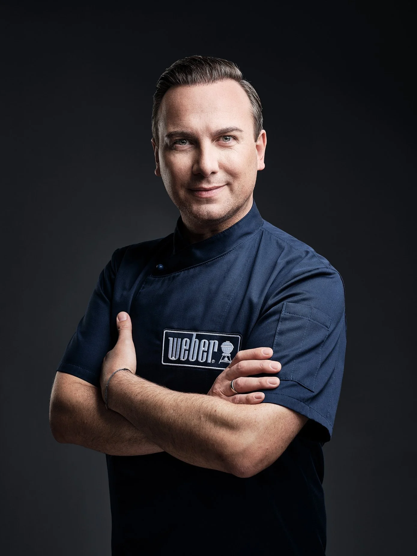Ein Mann in einem dunklen Hemd mit einem "Weber"-Logo, die Arme verschränkt.