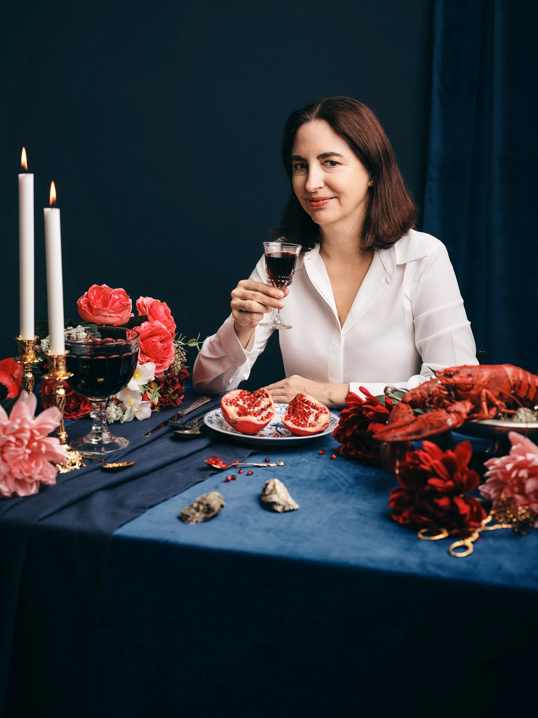 Frau sitzt an einem festlich gedeckten Tisch mit Kerzen, Blumen und Meeresfrüchten, hebt ein Glas Rotwein und lächelt.