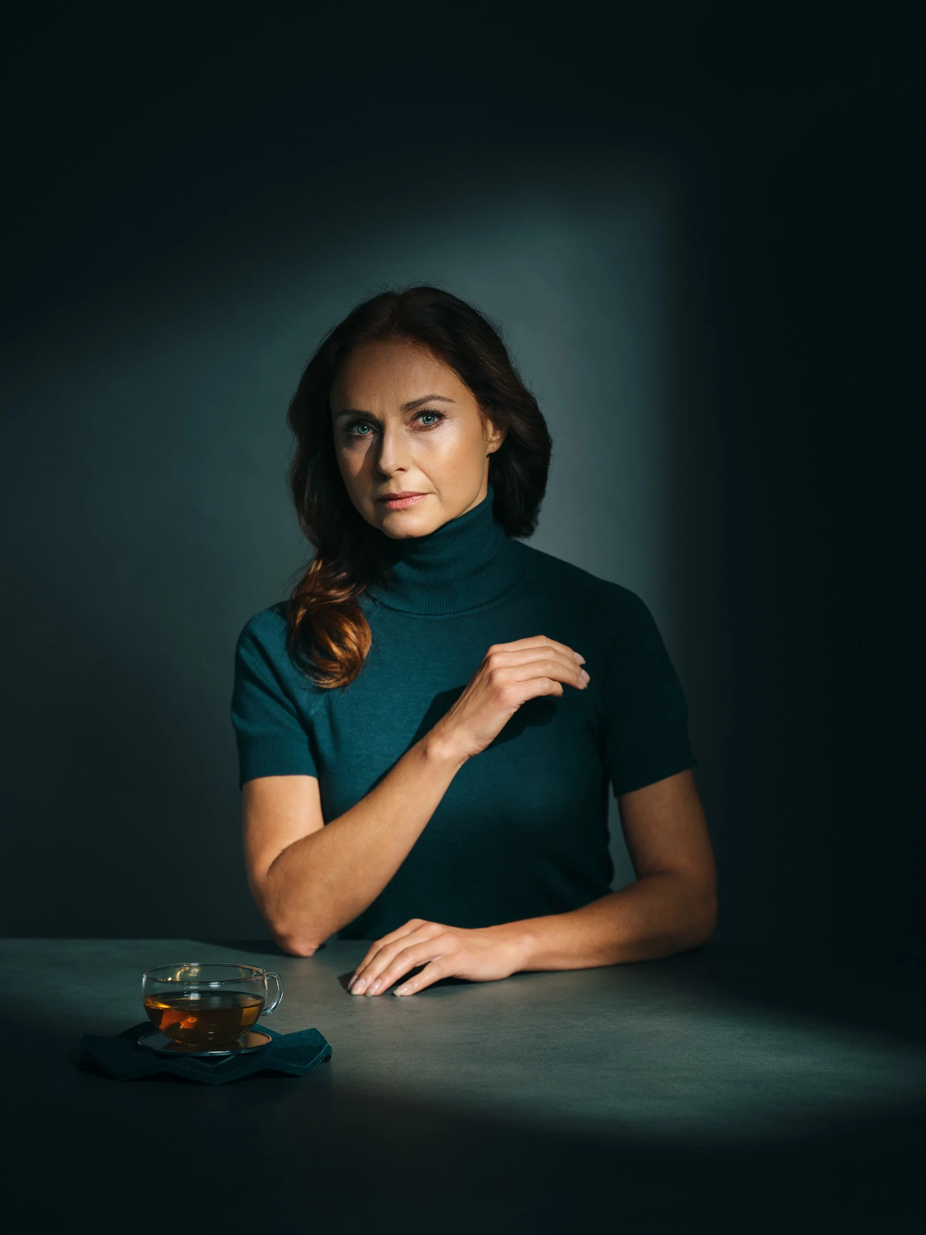 Eine Frau mit braunen, gewellten Haaren in einem blauen Rollkragenpullover sitzt an einem dunklen Tisch. Vor ihr steht eine Tasse Tee auf einer Unterlage. Der Hintergrund ist dunkel und schlicht.