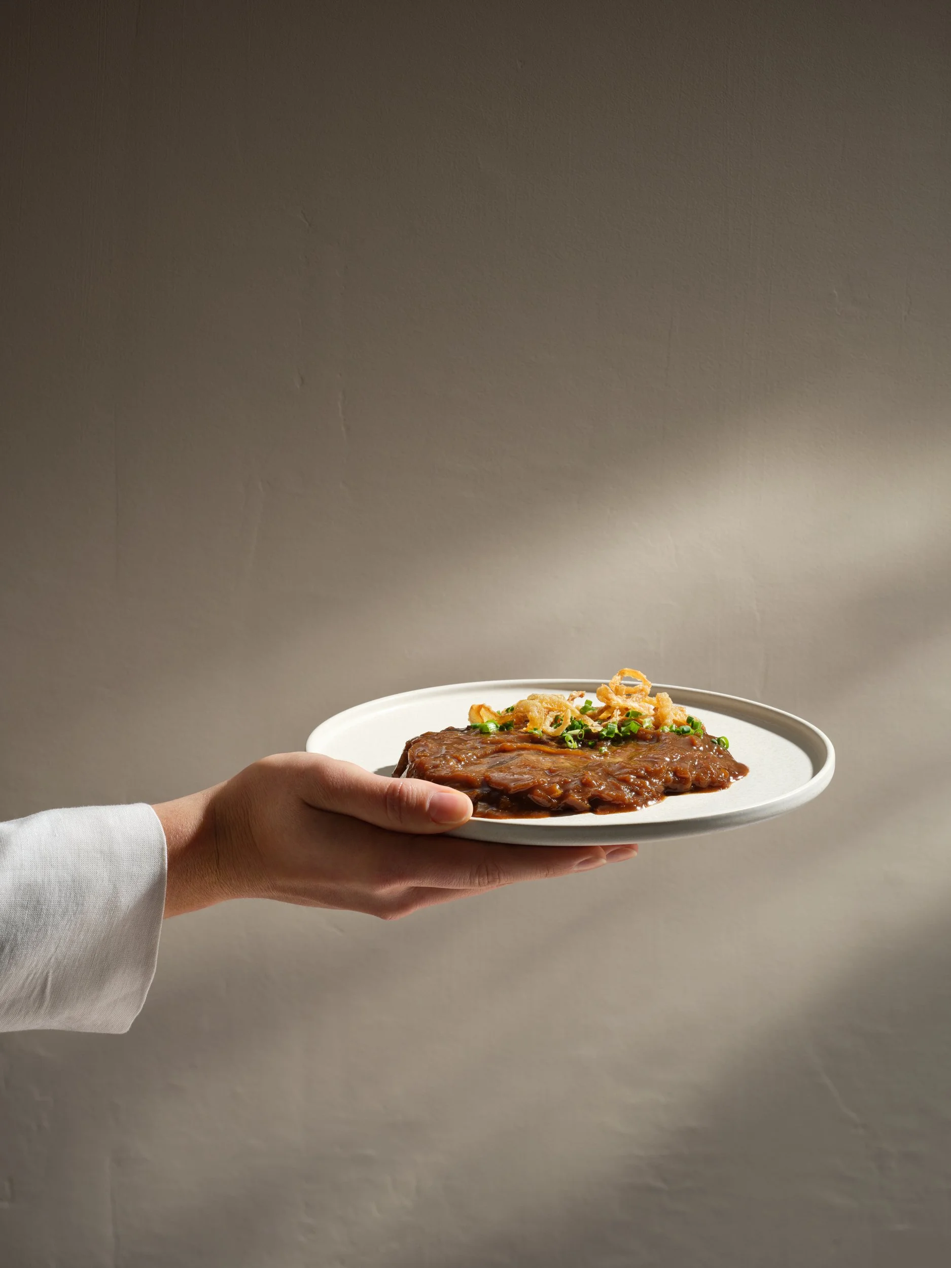 Eine Hand hält einen weißen Teller mit Fleisch in braicher Soße und Garnitur aus Frühlingszwiebeln und Zwiebelringen vor neutralem Wand.