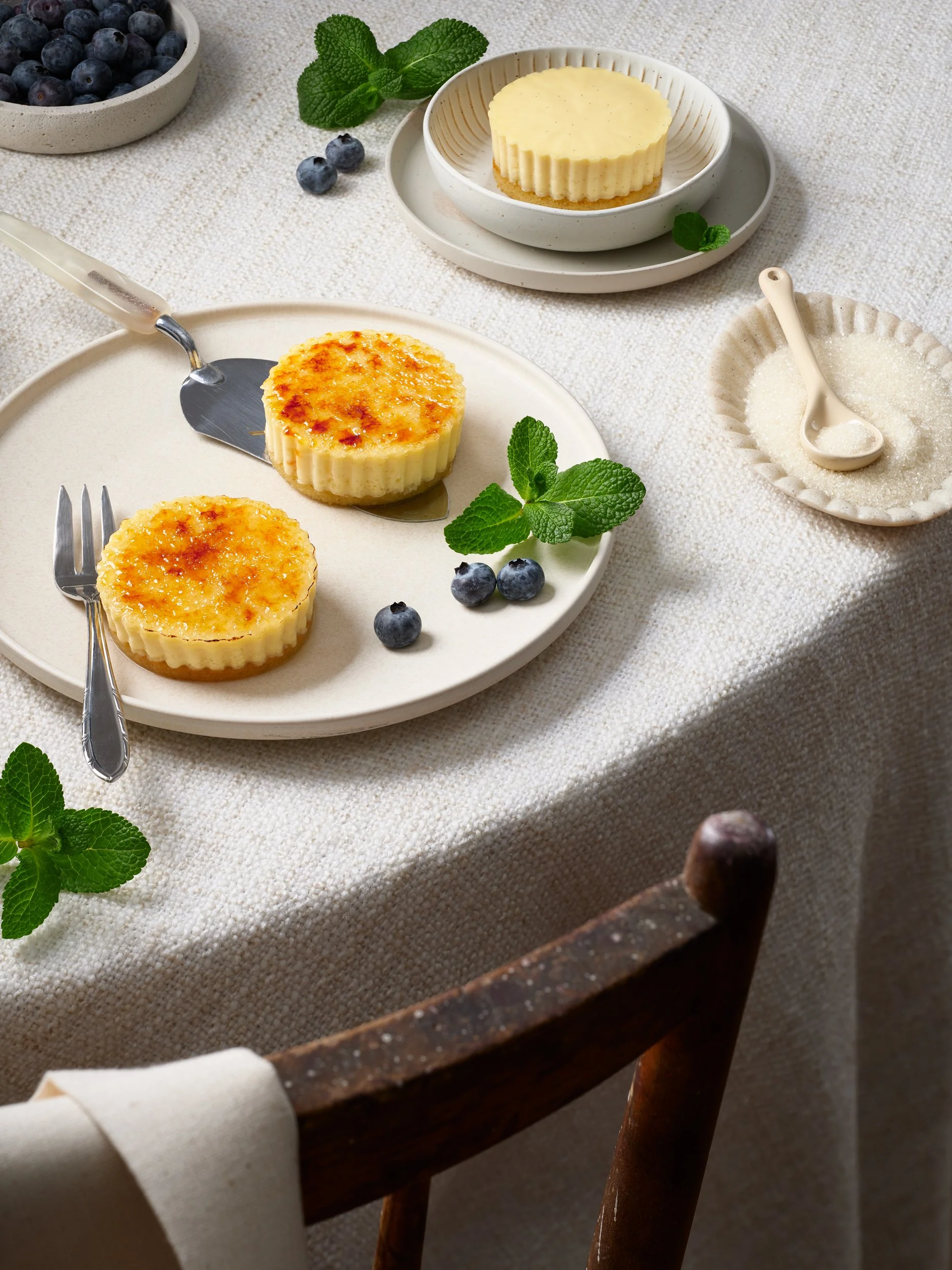 Zwei Käsekuchen auf einem weißen Teller, mit frischen Minzblättern und Heidelbeeren, auf einem Stofftisch, mit Salz und Zucker in kleinen Schalen, und einem weißen Tortenmesser.