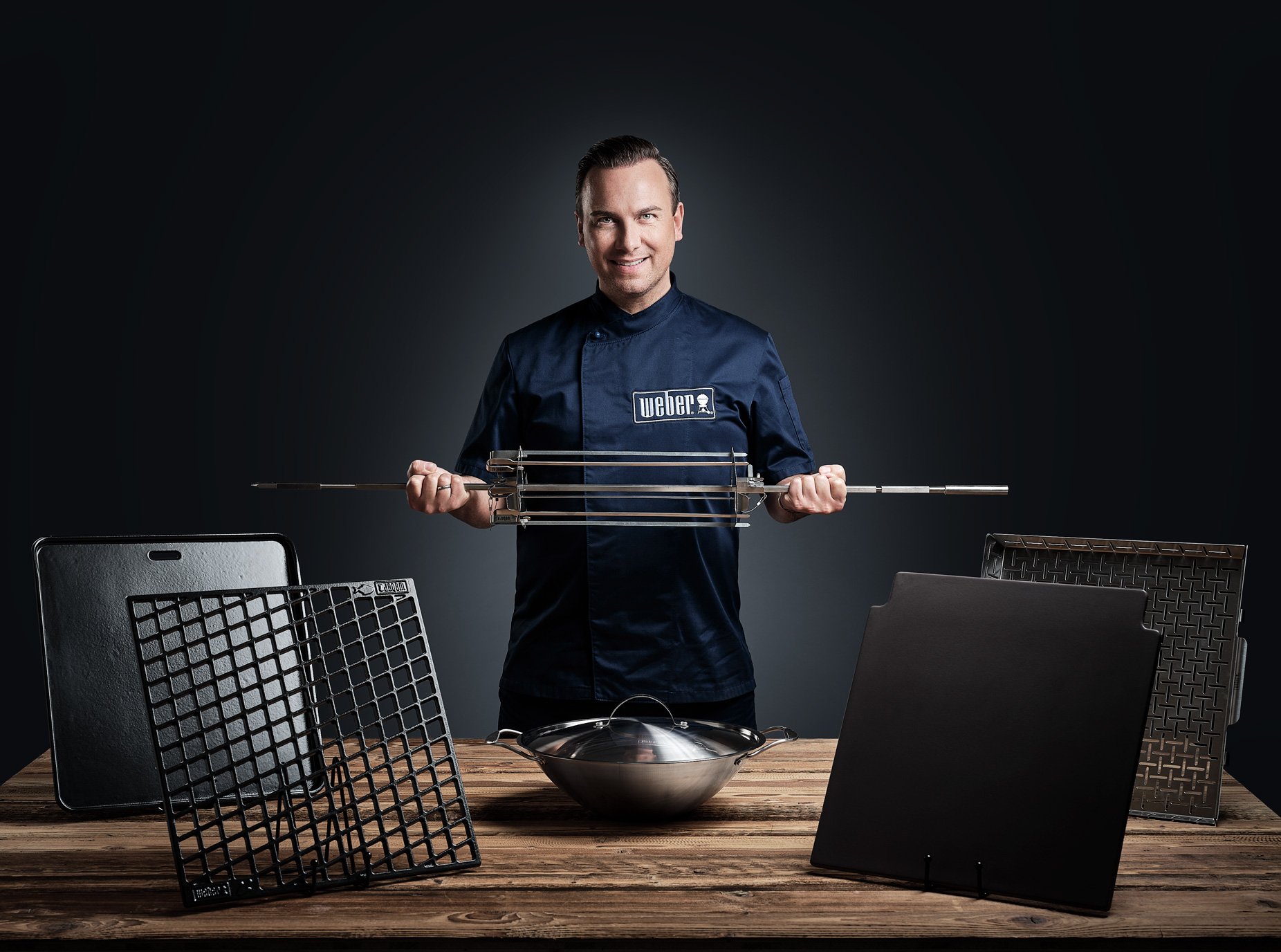 Person in Weber-Kleidung hält Grillspieß, umgeben von Grillzubehör, vor dunklem Hintergrund.