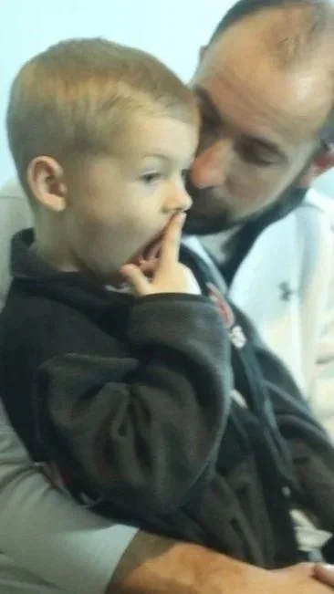 A man holding a young boy with red hair, who is placing his finger to his lips as his father whispers in his ear "after we're done we'll go for icedream".