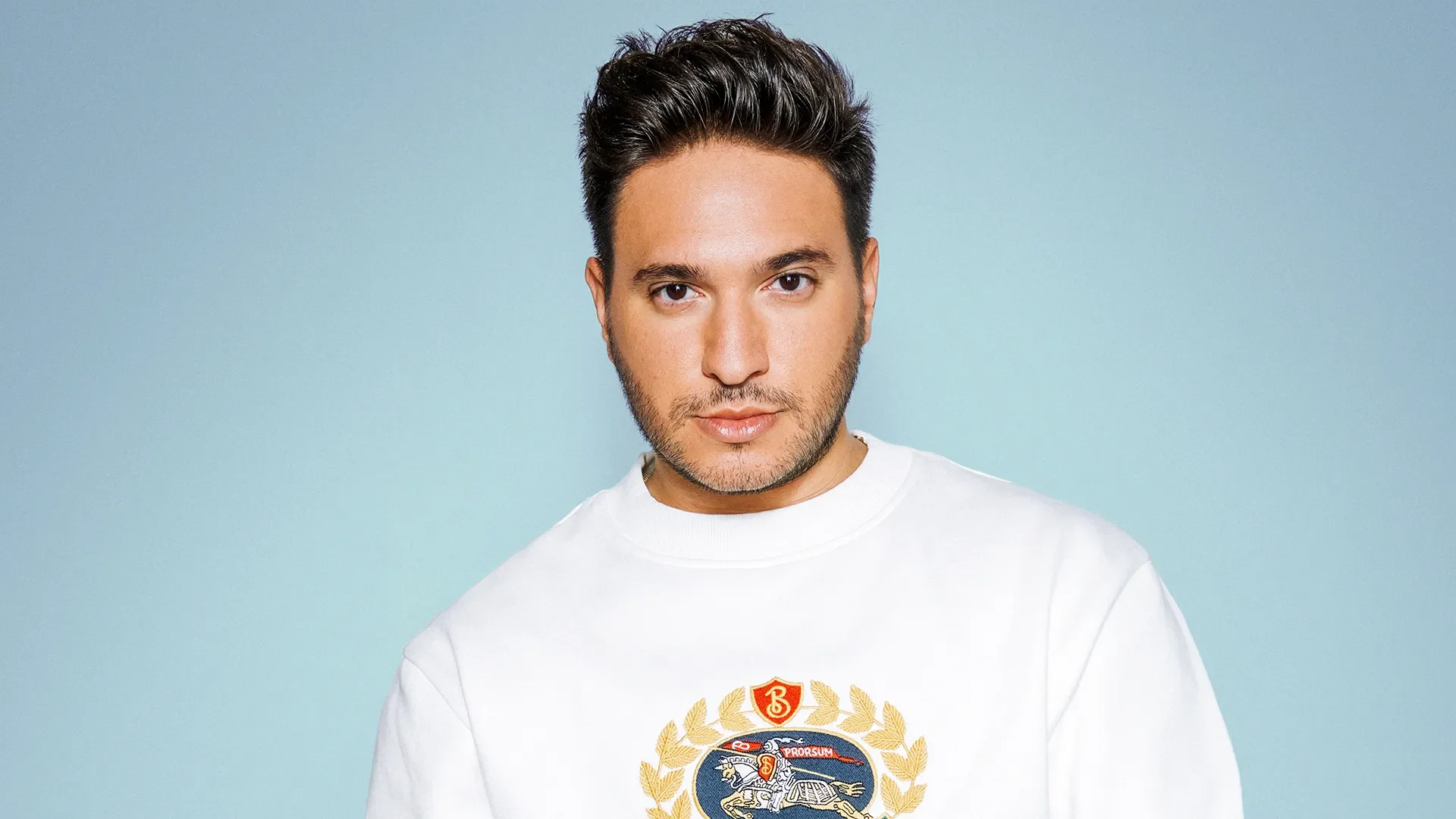 A man with dark hair styled upwards, light facial hair, wearing a white sweatshirt with a colorful crest and gold laurel design, against a light blue background.