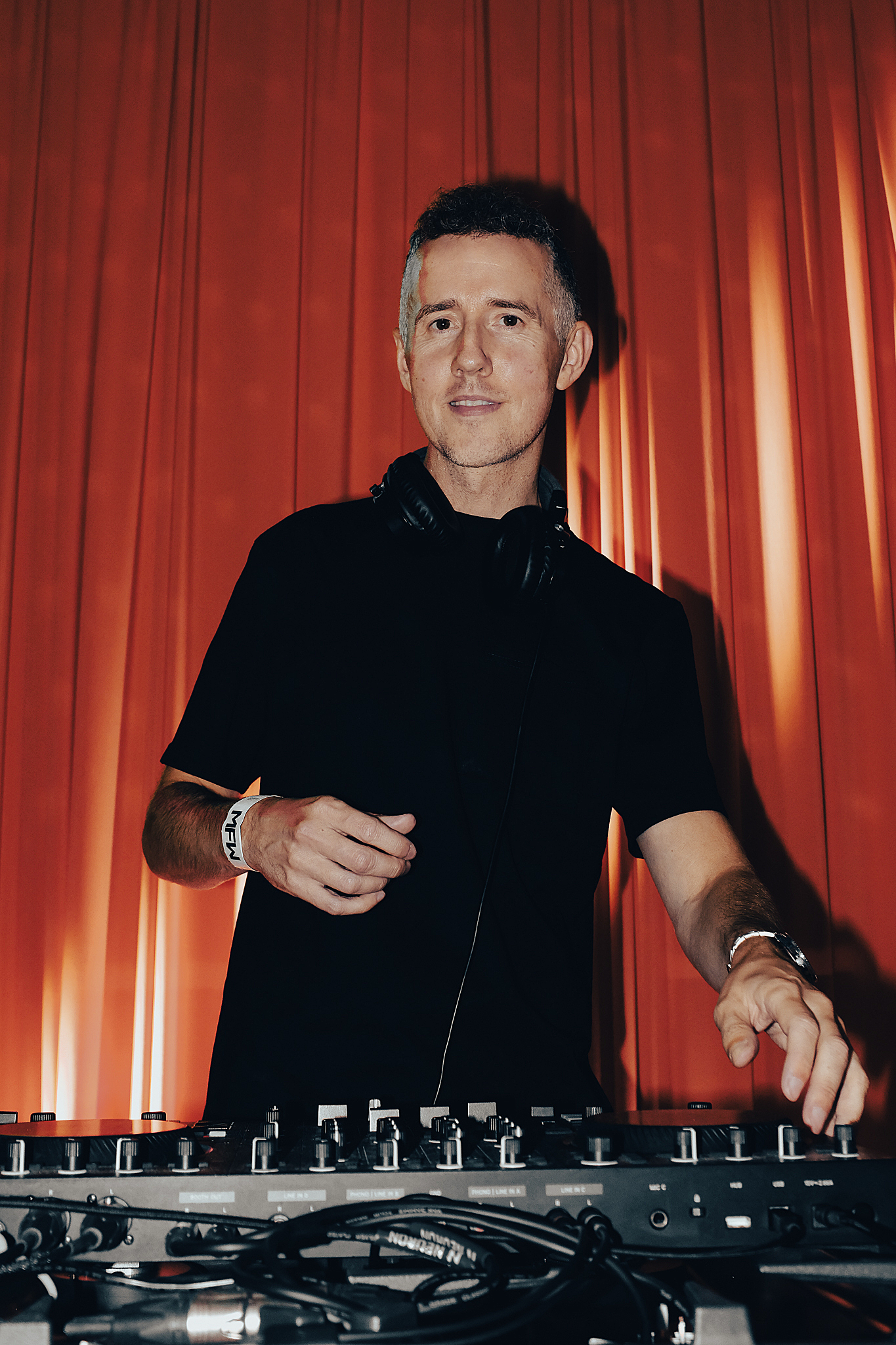 A male DJ wearing a black t-shirt and a white wristband with the word 'MEN' is standing behind a DJ mixer, adjusting controls with his right hand. He has a pair of headphones around his neck and is set against an orange curtain backdrop.