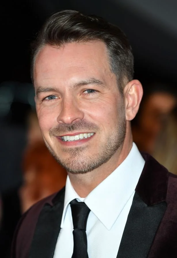 A smiling man with short, styled hair and a trimmed beard, wearing a tuxedo with a black tie, at a formal event.