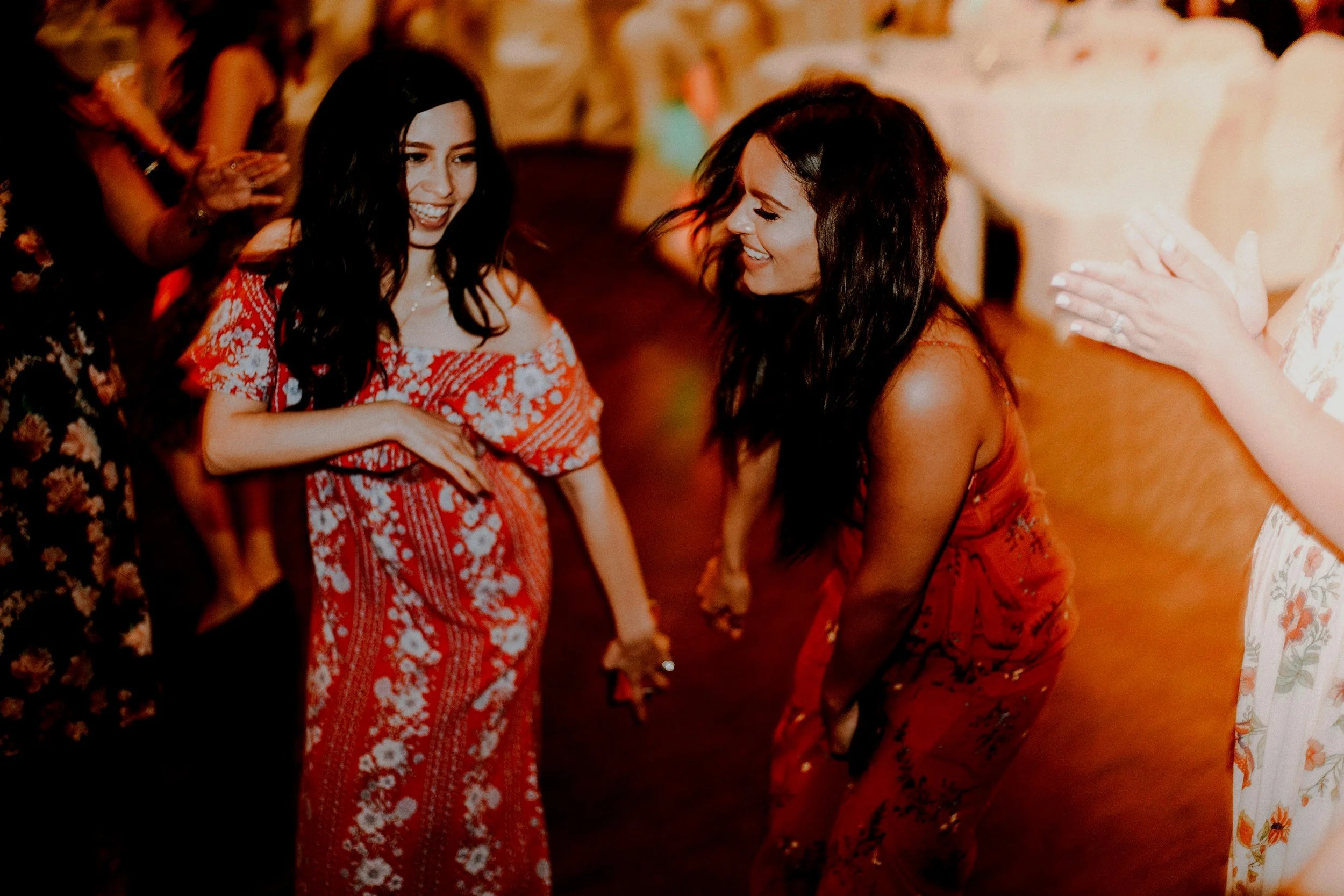Two women in red dresses are holding hands, smiling, and dancing together at a party.