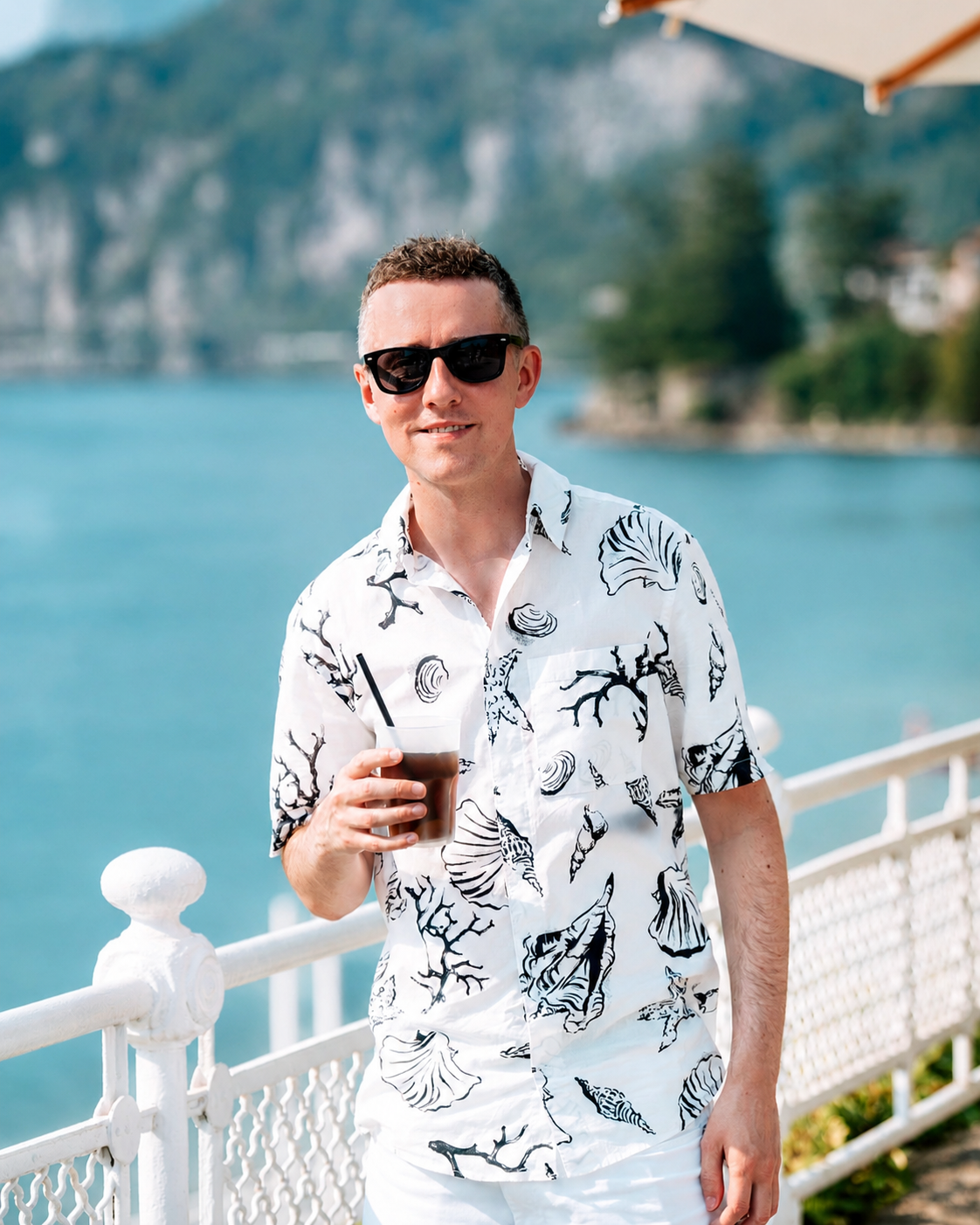 Man wearing sunglasses and a white short-sleeved shirt with black tropical leaf and shell patterns, holding a glass of iced coffee with a straw, standing outdoors near a white railing with water and greenery in the background.