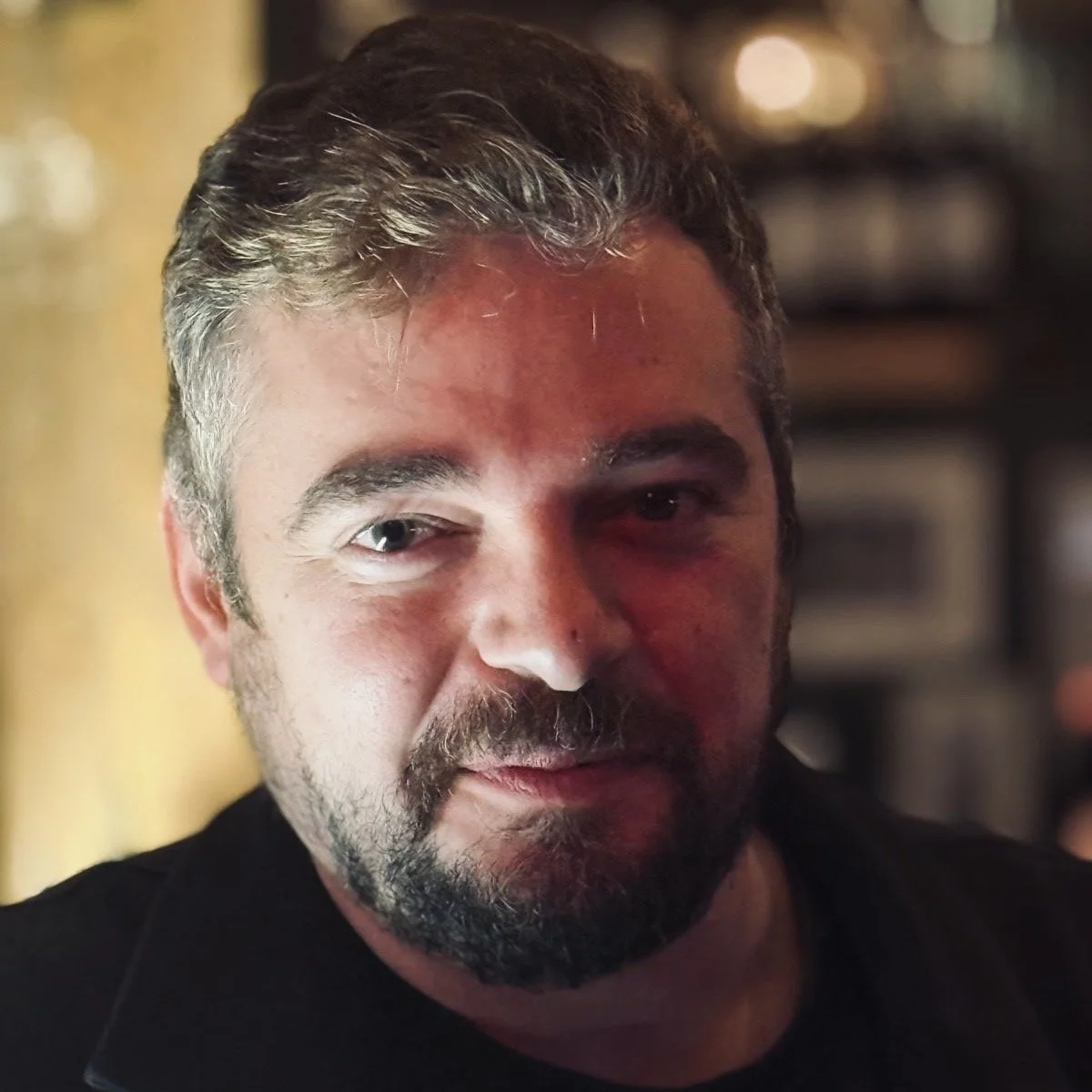 A close-up of a man with gray hair and a beard, looking at the camera in a dimly lit indoor setting.