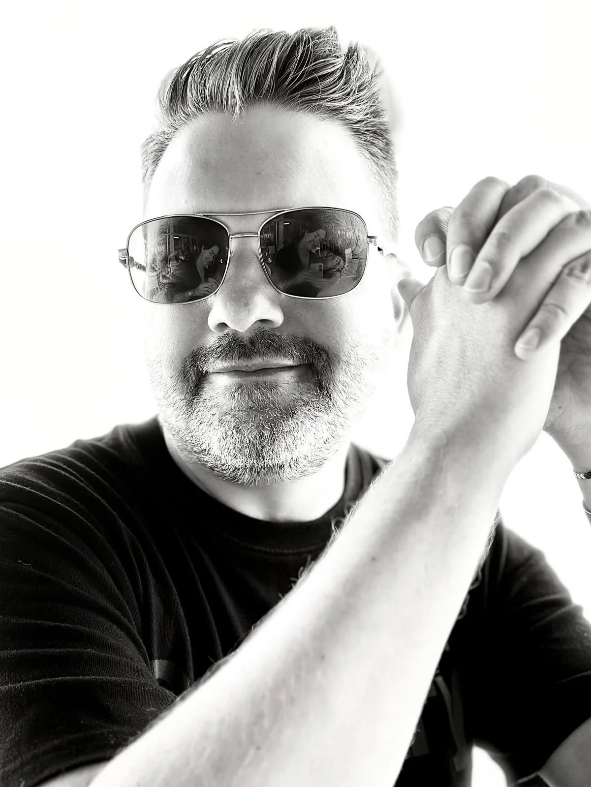 Black and white close-up portrait of a man with sunglasses, a beard, and styled hair, posing with his hands clasped together.