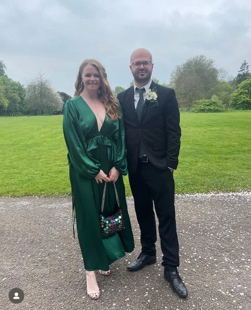 A woman in a long green dress and a man in a black suit standing on a grassy outdoor area with trees and a cloudy sky in the background.