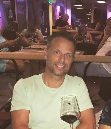 A man smiling and holding a glass of red wine in a restaurant or bar with other people dining in the background.