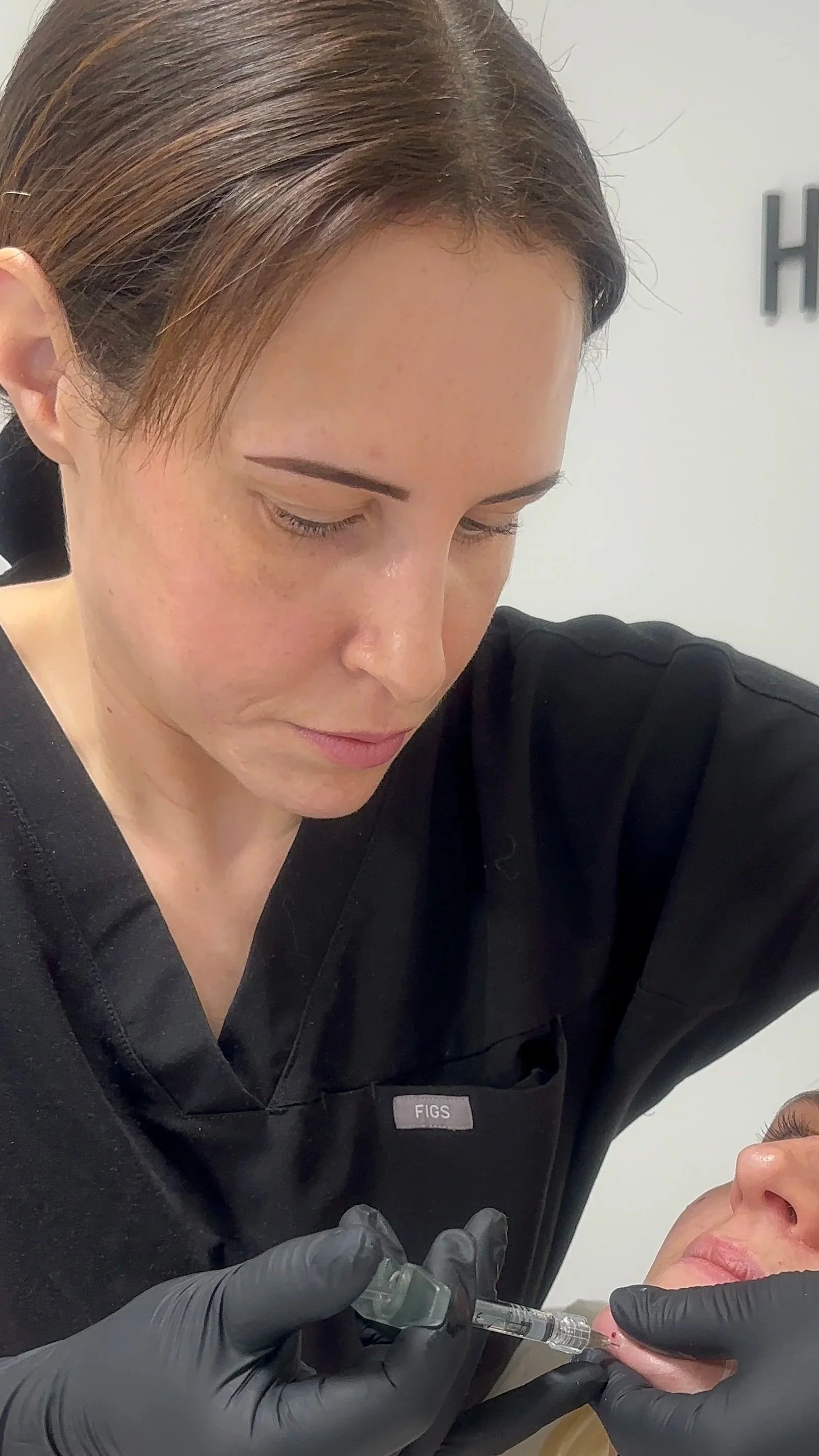 A woman in black scrubs and black gloves performs a facial injection on a patient who is lying down. The woman has brown hair and is focusing on the procedure.