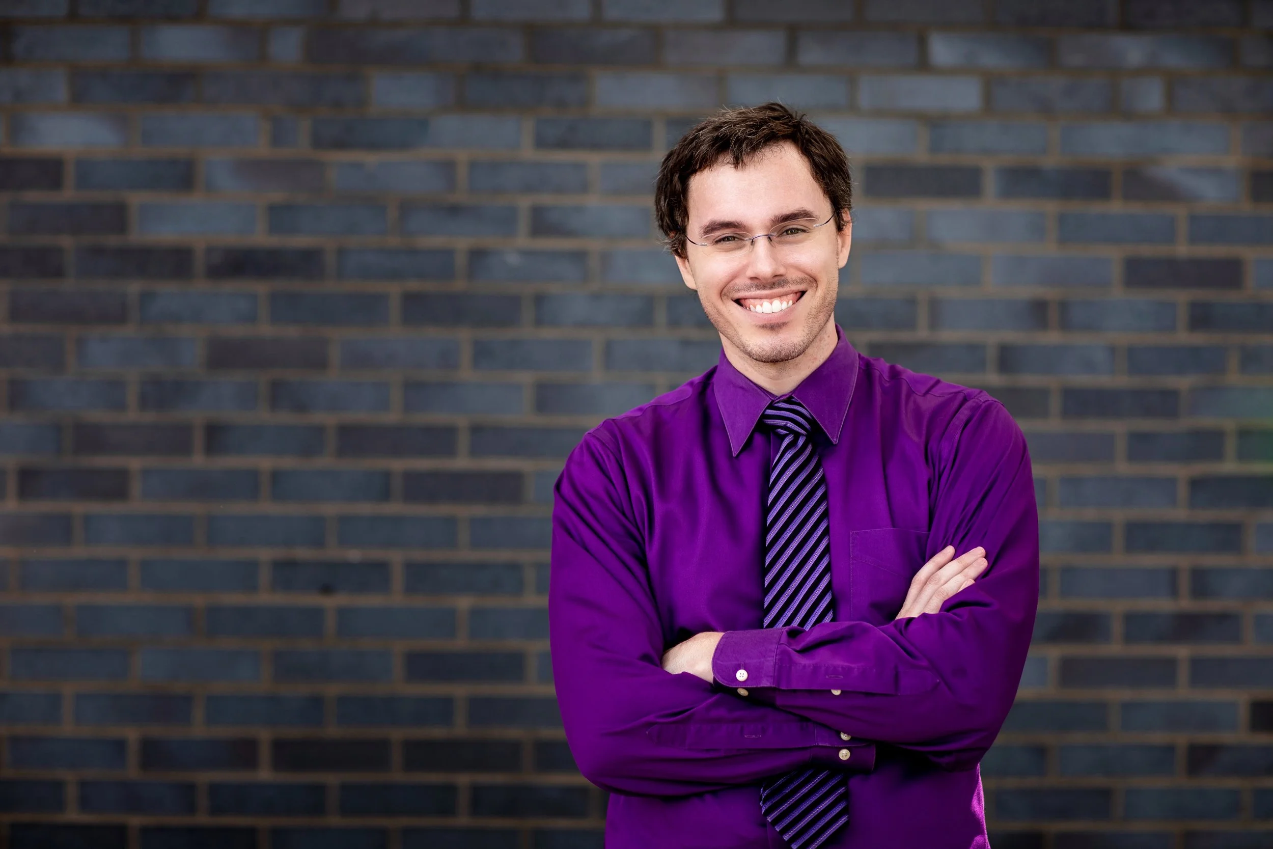 A young man with brown hair, glasses, and a smile, standing with arms crossed in front of a brick wall. He is wearing a purple shirt and a matching striped purple and black tie.