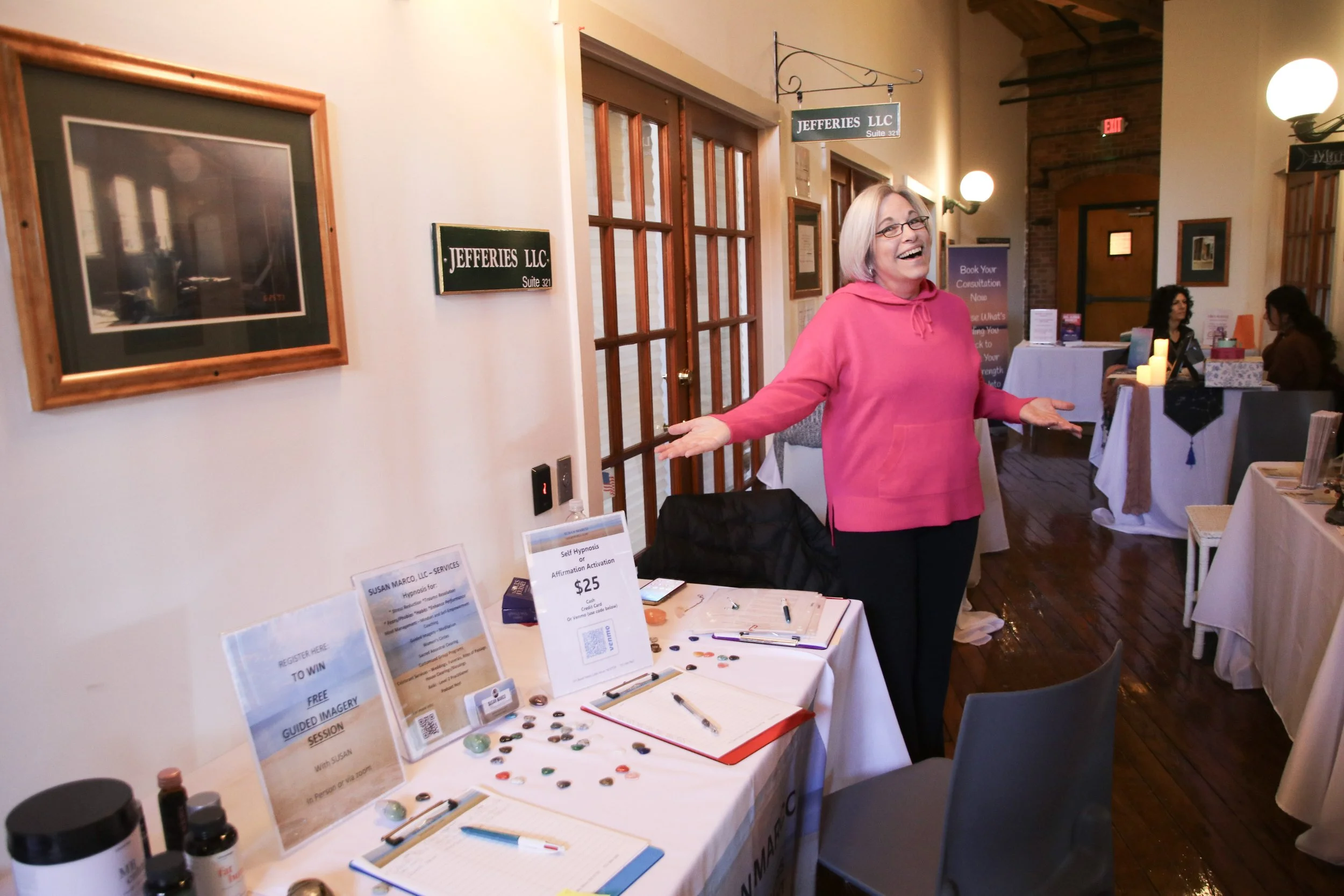 A woman in a pink hoodie standing at a table with various items, smiling and gesturing with her arms extended, in a room with tables, decorations, and other people.
