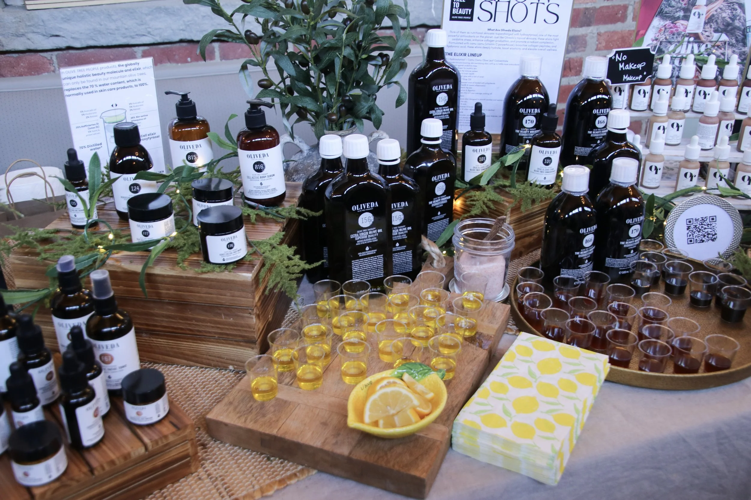 Display of skincare and wellness products, including bottles of oils and lotions, on a table decorated with greenery, with shot glasses filled with yellow and dark liquids, a bowl of lemon slices, and informational posters in the background.