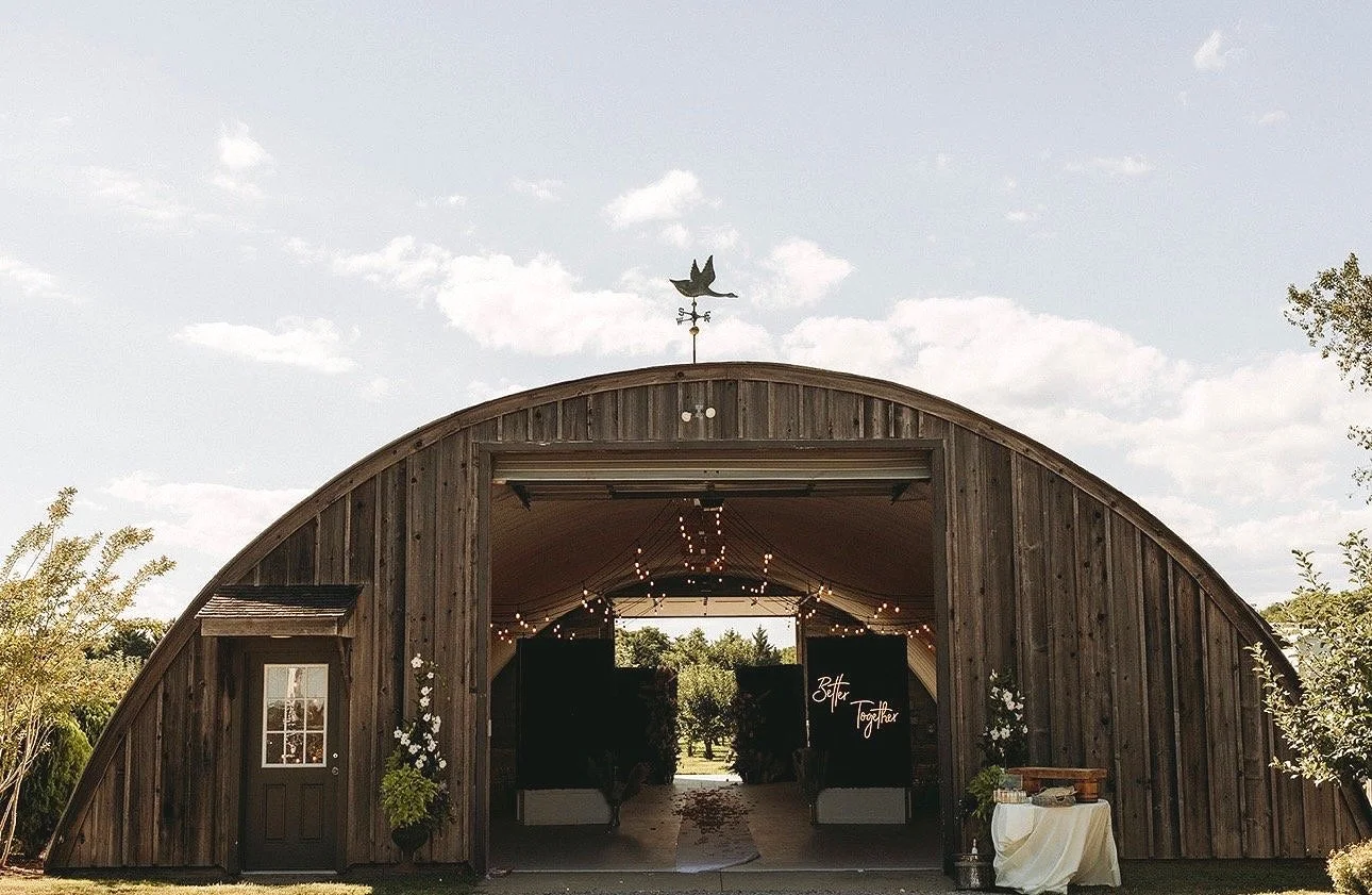 A rustic wooden barn with string lights hanging inside, decorated with flowers and a sign that reads 'Better Together,' likely set up for a wedding celebration.