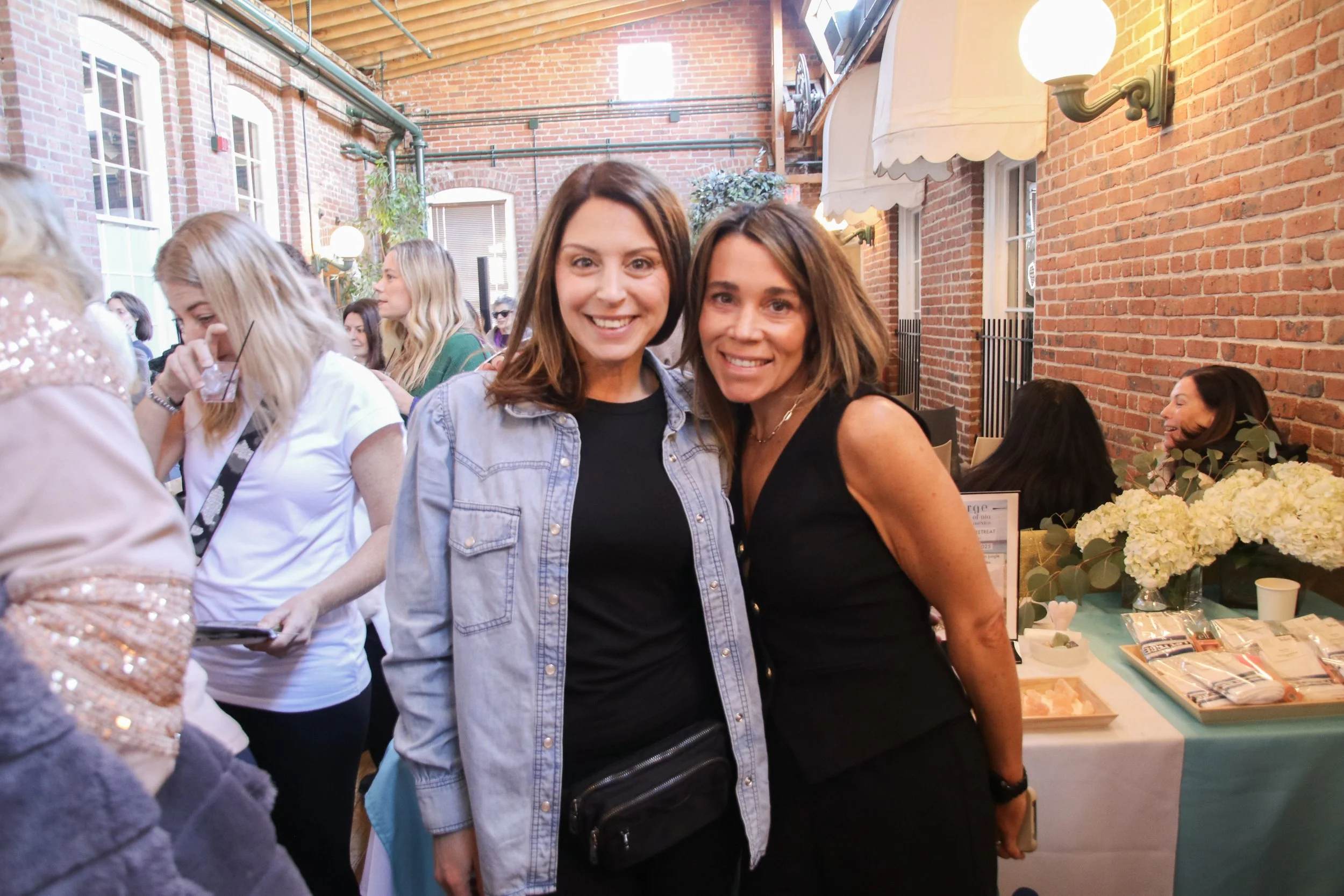 Two women smiling and posing for a photo at an indoor social event, with people and a table of flowers and items in the background.