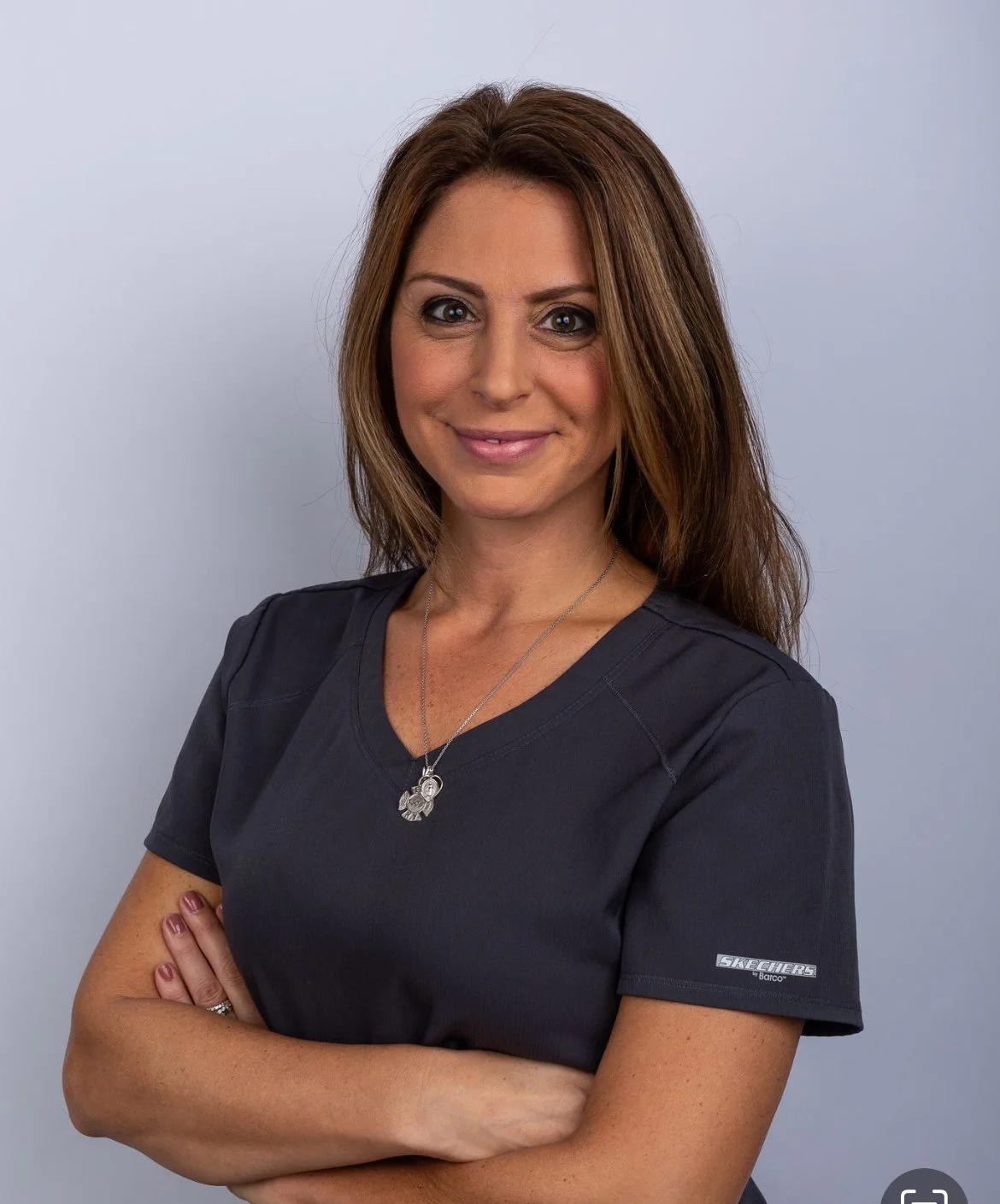 A woman with shoulder-length brown hair, wearing a dark gray Scrubs shirt and silver jewelry, stands with her arms crossed against a light gray background, smiling at the camera.