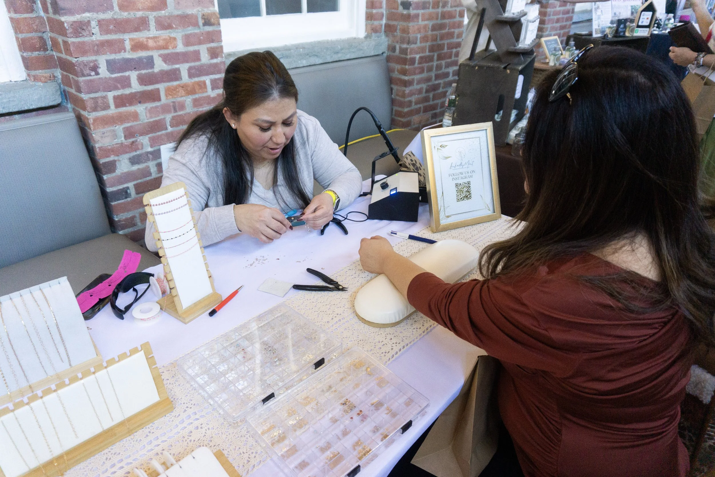 Two women seated at a jewelry table, one is making jewelry with tools while the other displays a jewelry piece, with display stands, tools, and a sign with a QR code on the table.
