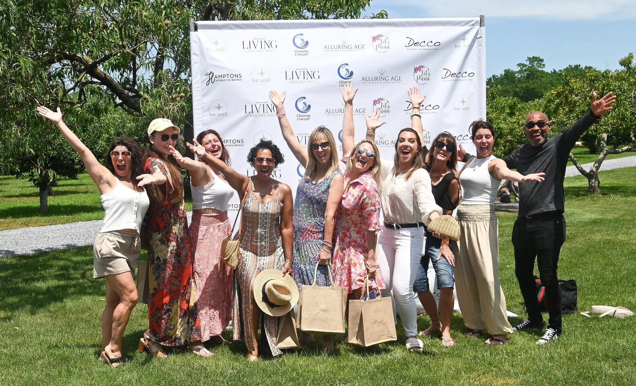 A group of people smiling and celebrating outdoors in front of a branded backdrop, on a sunny day with green trees and grass.