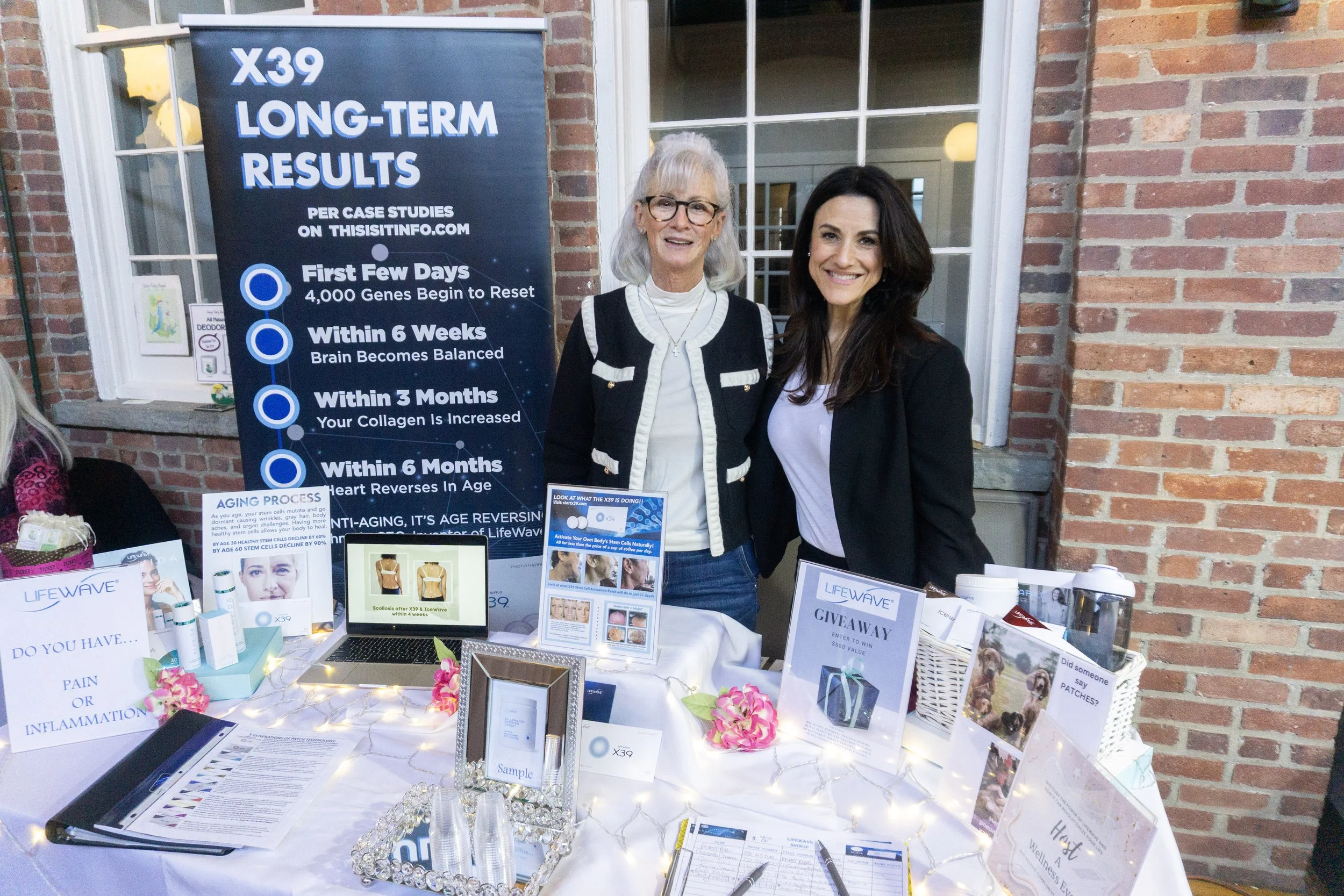 Two women are standing behind a table at a booth promoting LifeWave X39, a health and wellness product. The booth features pamphlets, a laptop, and signage about long-term results of X39. The background includes a brick wall and a large poster about 
