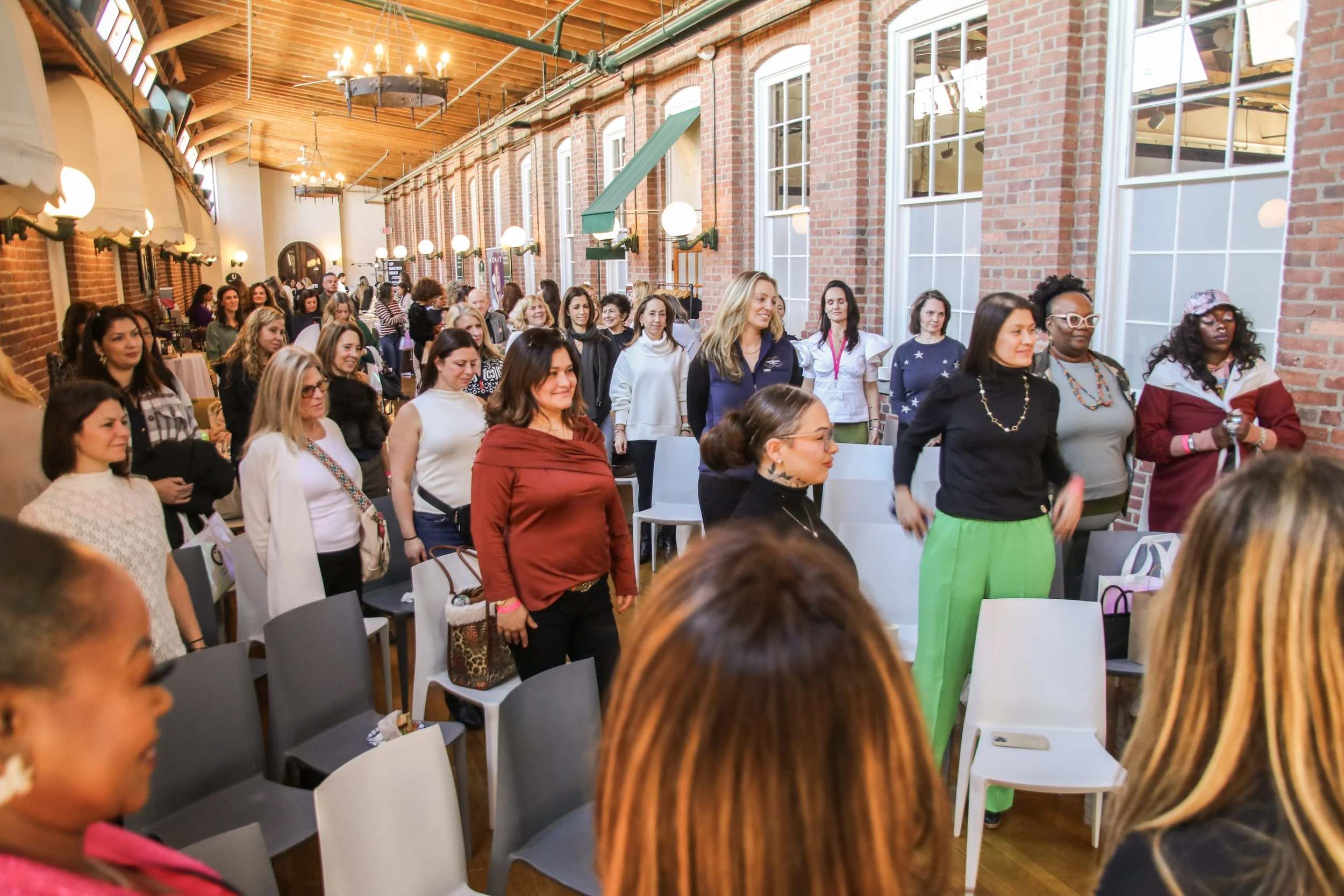 A group of women standing in a spacious, well-lit room with brick walls, large windows, and wooden ceiling beams, participating in an indoor event or seminar.