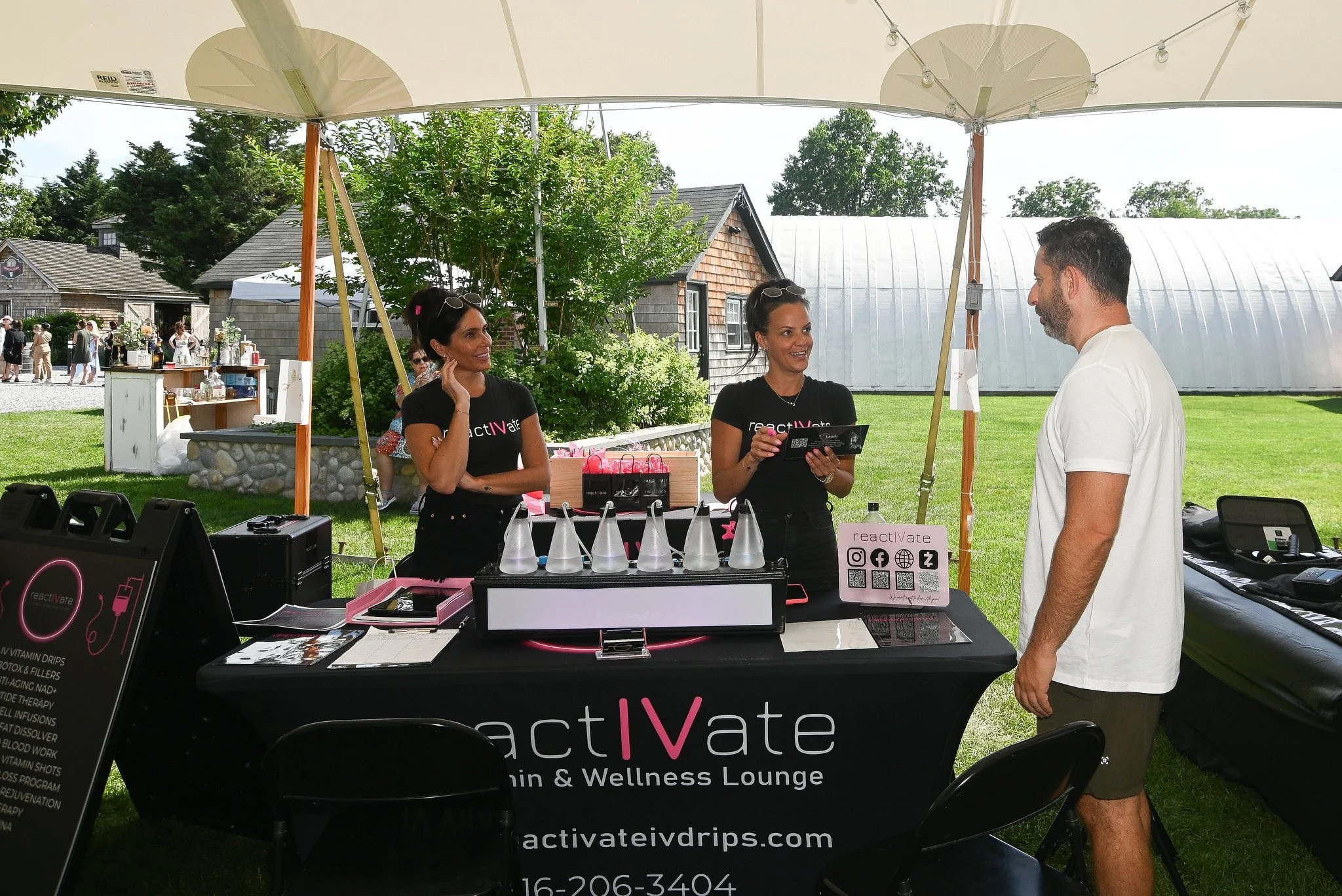 Two women and a man at a reactivAte vitamin drip booth outdoors, under a white canopy, with a grassy field and small buildings in the background.