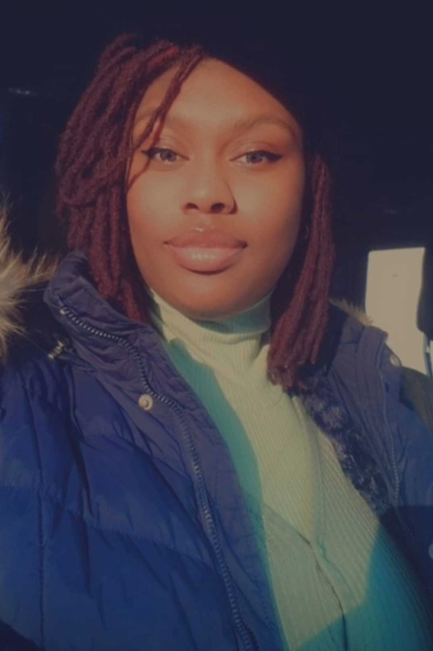 A woman with reddish-brown dreadlocks wearing a navy blue winter jacket and a light yellow turtleneck sweater, smiling outdoors at night.