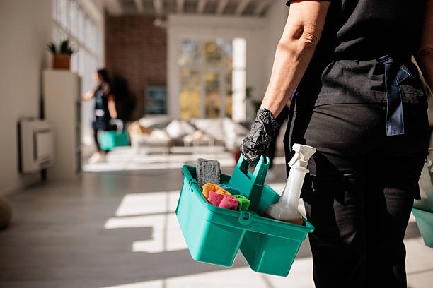 Person holding a cleaning caddy with spray bottles and towels in a bright living room.