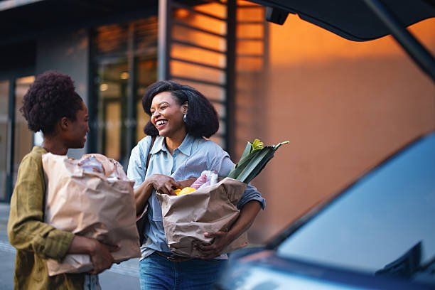 Two women exchanging grocery bags outside at sunset, smiling and chatting.