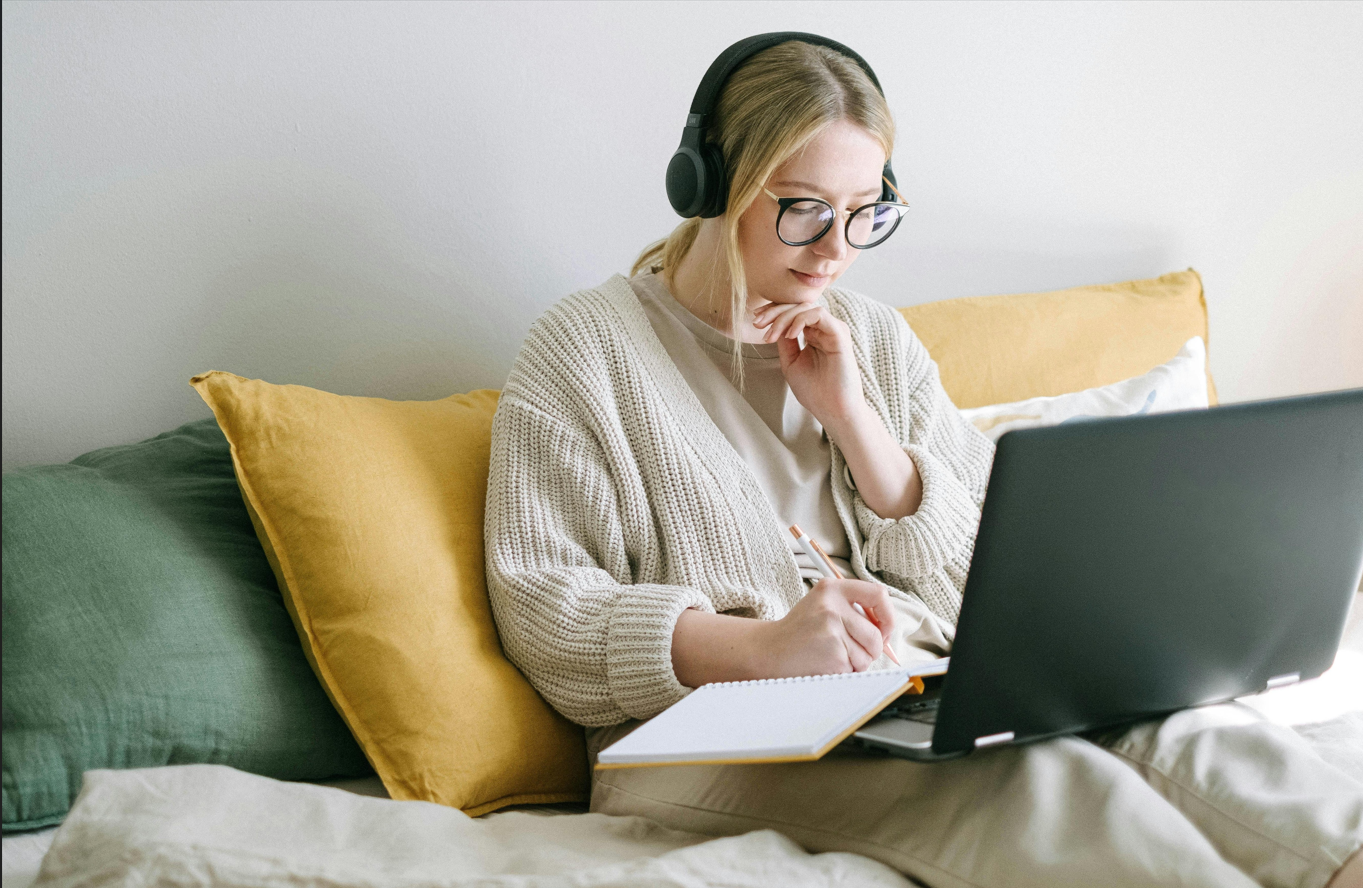 A woman with glasses and headphones sitting on a bed with yellow and green pillows, working on a laptop and writing in a notebook.