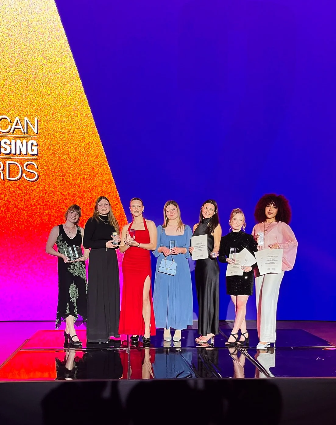 Seven women standing on a stage, holding awards and certificates, at an event with colorful lighting and background.