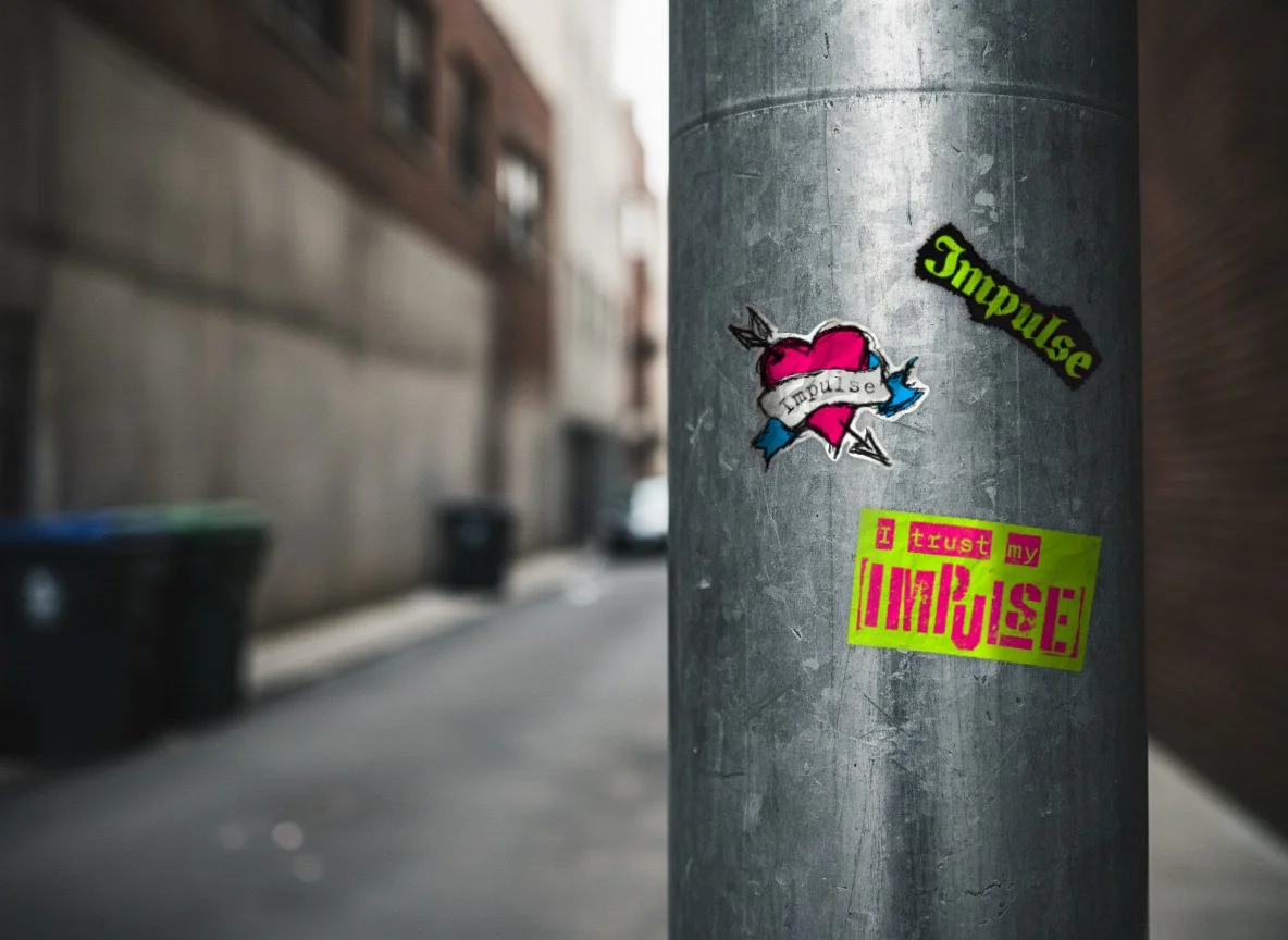 Street scene with graffiti stickers on a metal pole, including a pink heart with a banner reading "Impulse," a sticker with green and black text "Impulse," and a pink and green sticker reading "I trust my IMPULSE!"