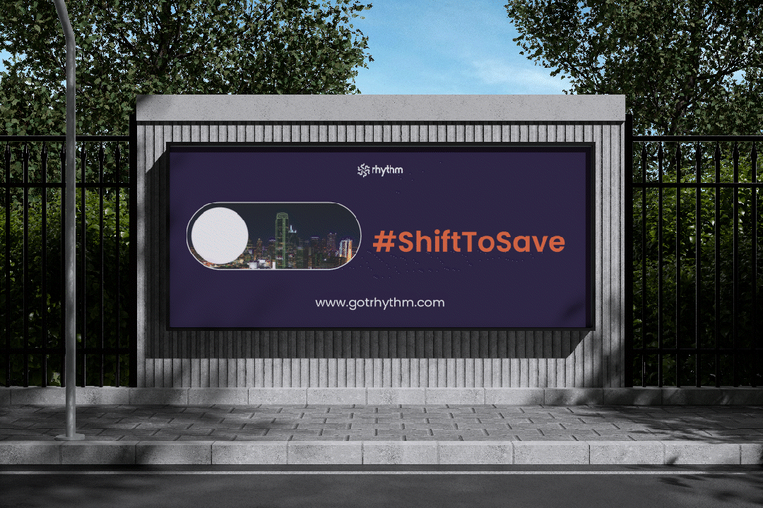 Digital billboard on a sidewalk fence displaying logo of rhythm and hashtag #ShiftToSave with a city skyline in background.