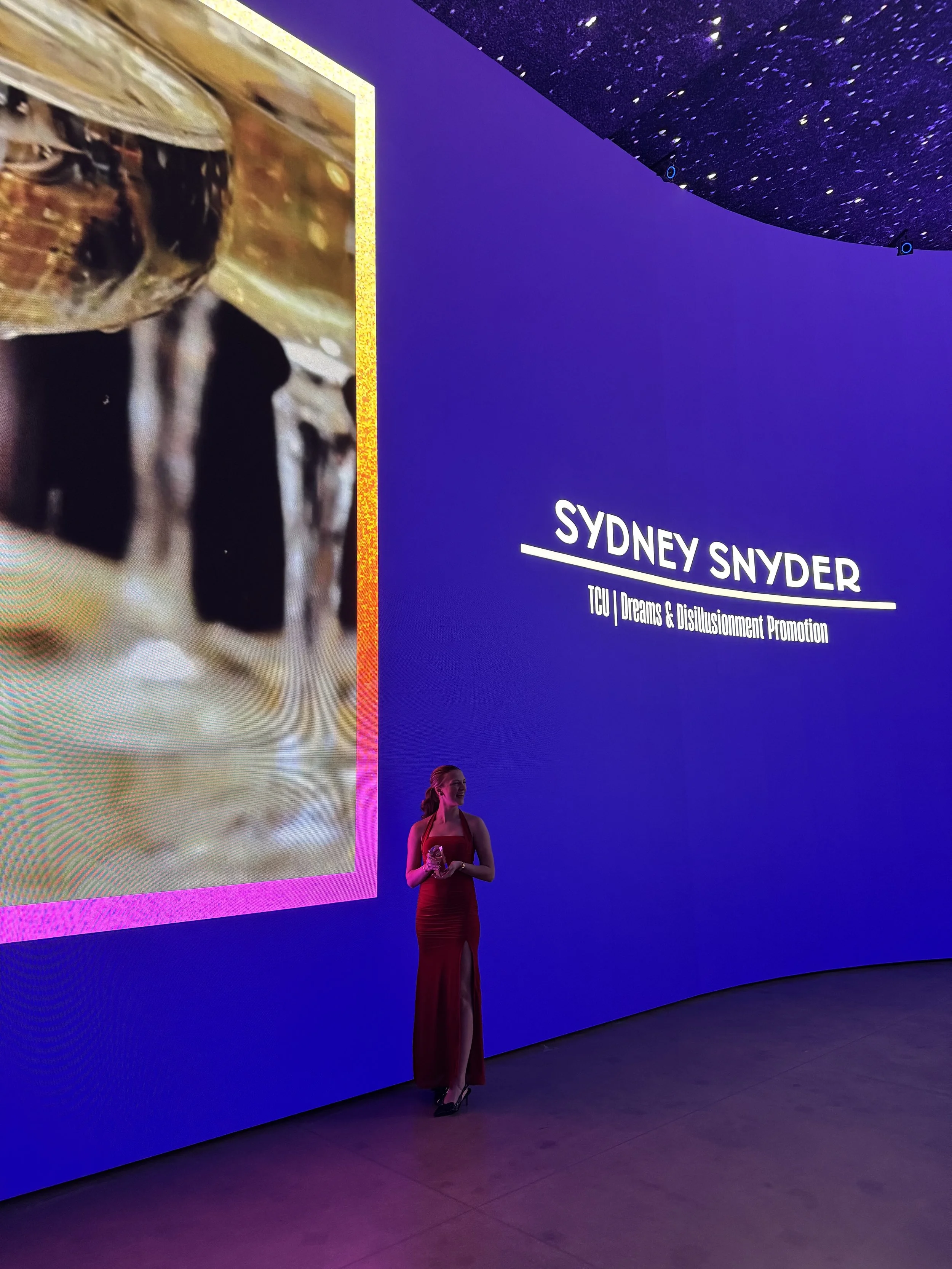 A woman in a red dress standing in front of a large blue screen with white text displaying "Sydney Snyder" and "TCU | Dreams & Disillusionment Promotion." The woman is holding an award or trophy.