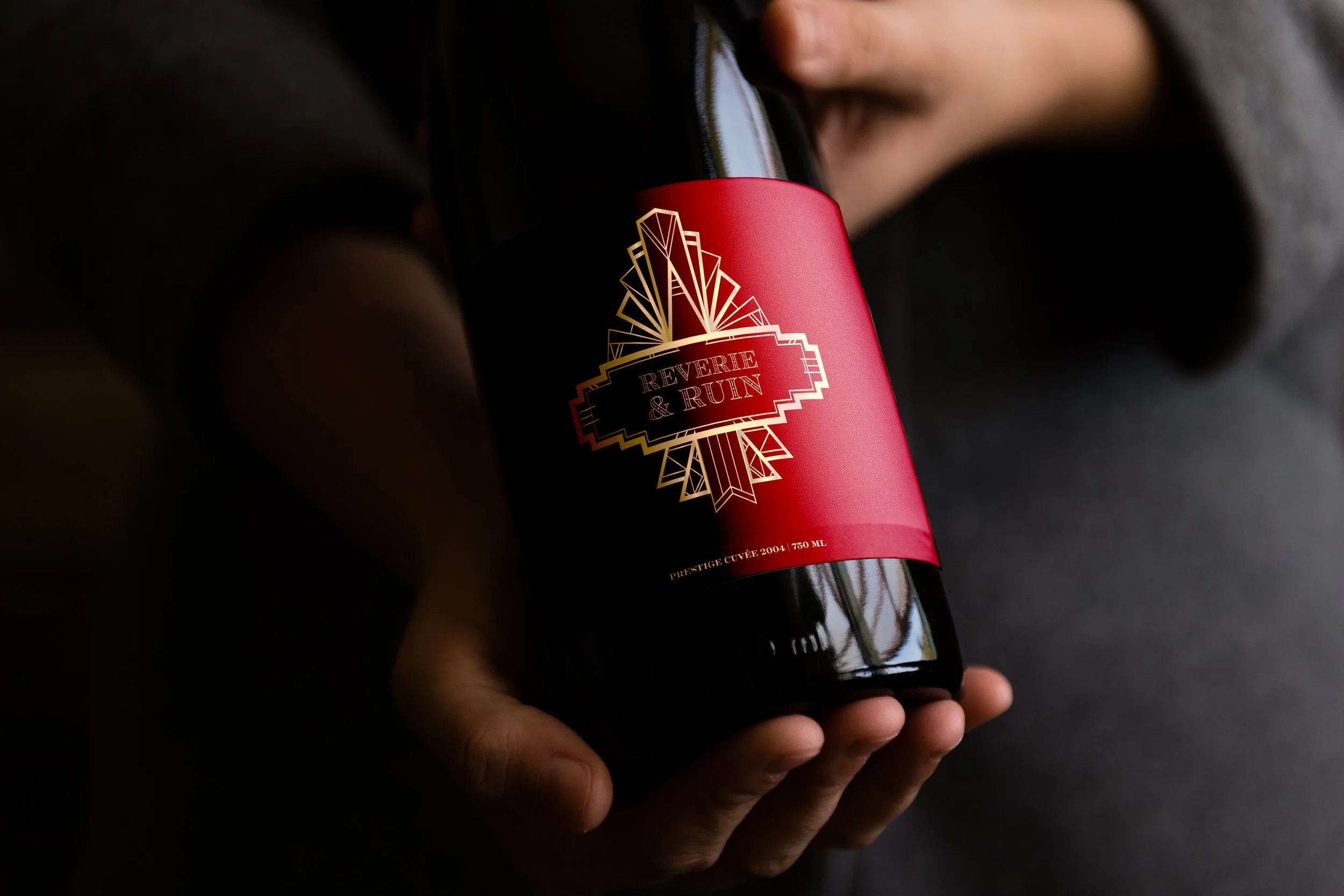 A person holding a black wine bottle with a red and gold label that says 'Reverie & Ruin' and features an art deco style design.