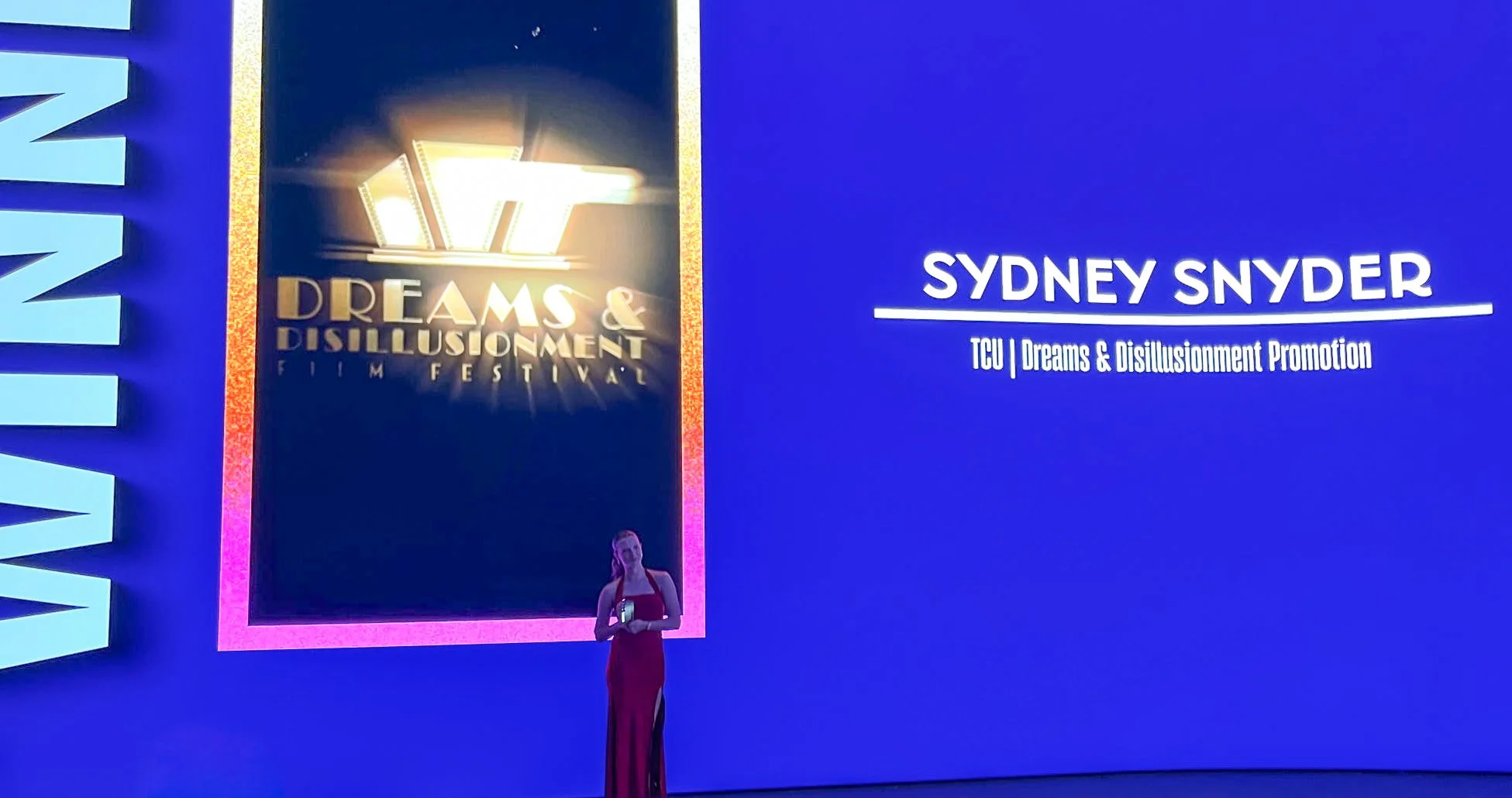 A stage presenting Sydney Snyder from TCU at the Dreams & Disillusionment Film Festival, with a woman in a red dress standing in front of a large blue screen.