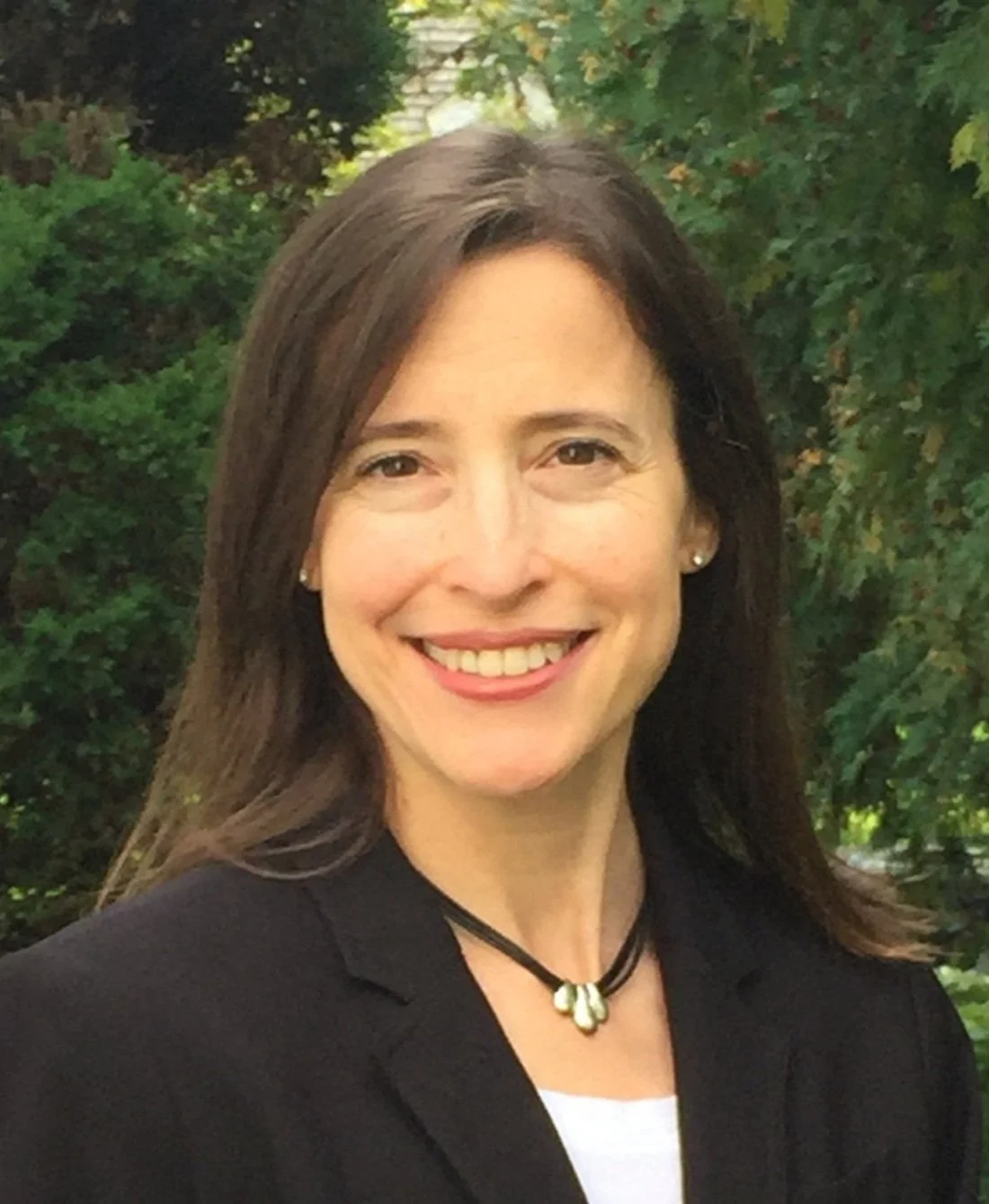 A woman with brown hair smiling outdoors, wearing a black blazer and a black necklace with a silver pendant.