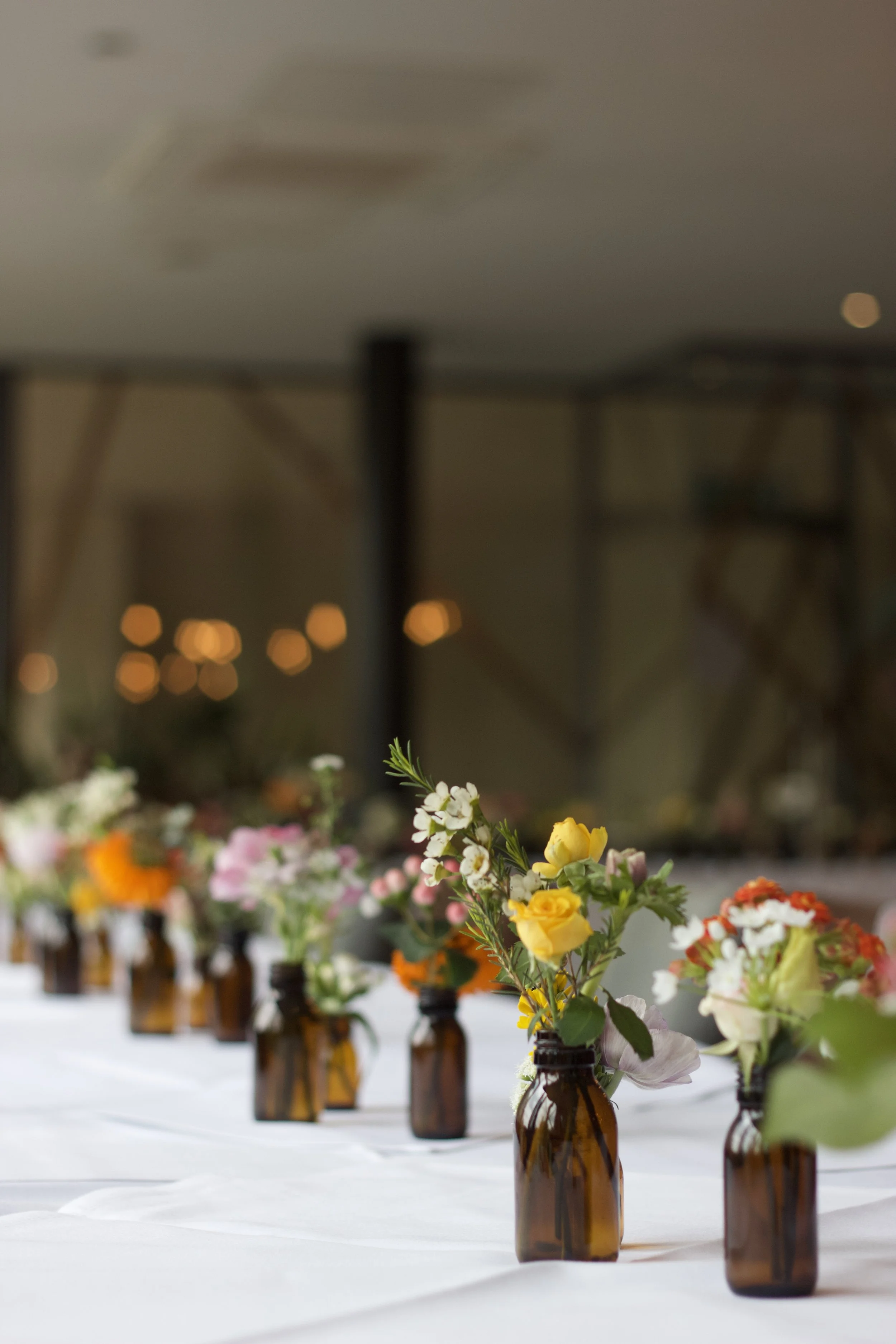 Small brown glass bottles with colorful flowers arranged inside, placed on a white tablecloth, with a blurred background.