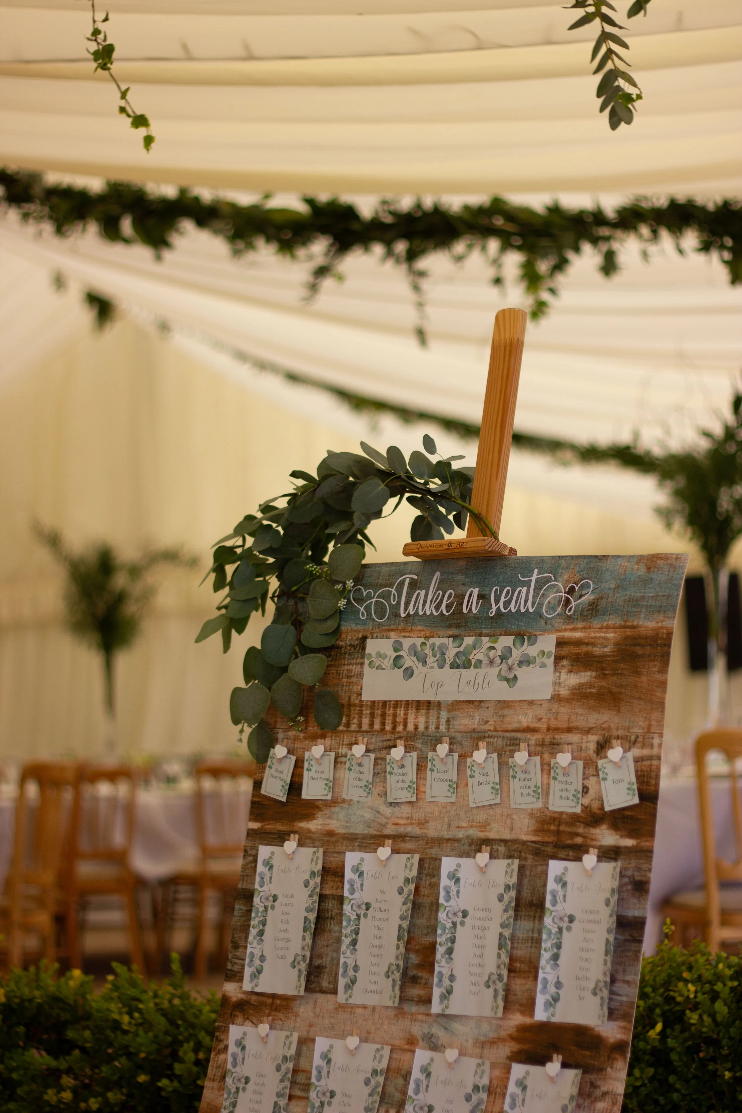 A wooden event seating chart with greenery decorations and a 'Take a seat' sign, displaying tables and guest names in a decorated venue.