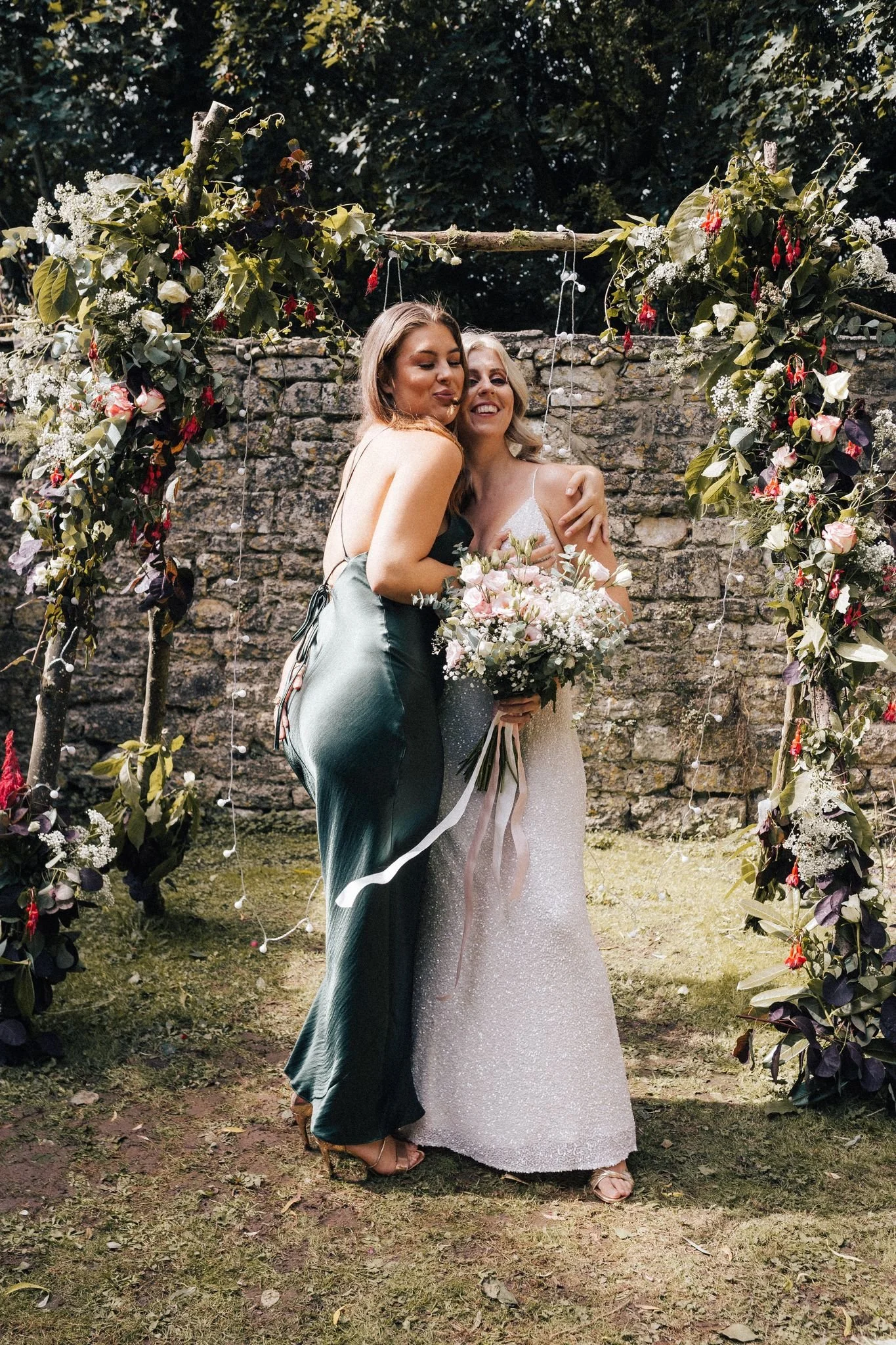 Two women, one in a white wedding dress and the other in a dark green gown, embracing and smiling outdoors in front of a floral arch during a wedding celebration.