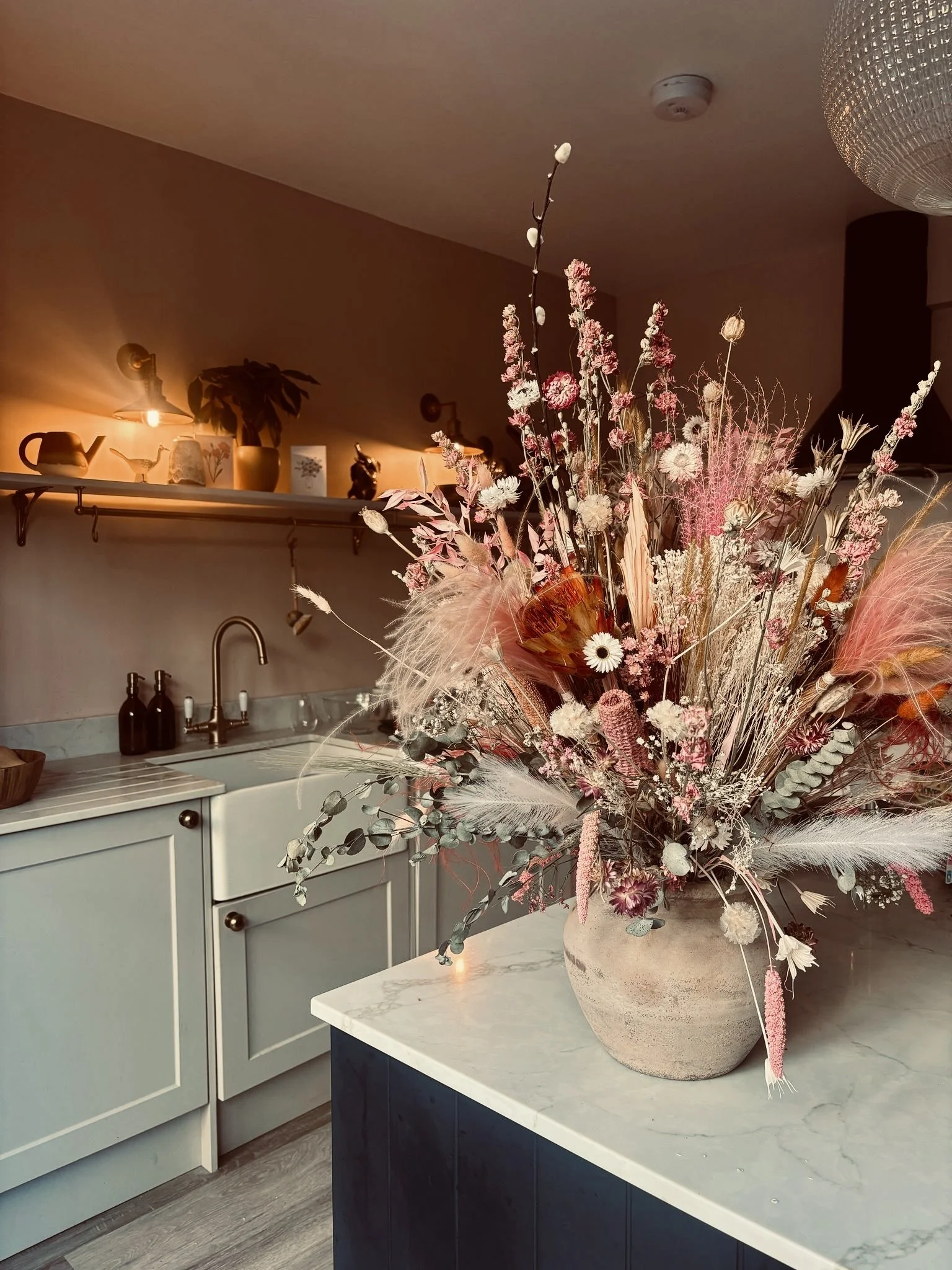 A large decorative flower arrangement with pink, white, and beige dried flowers in a beige pot on a kitchen counter.