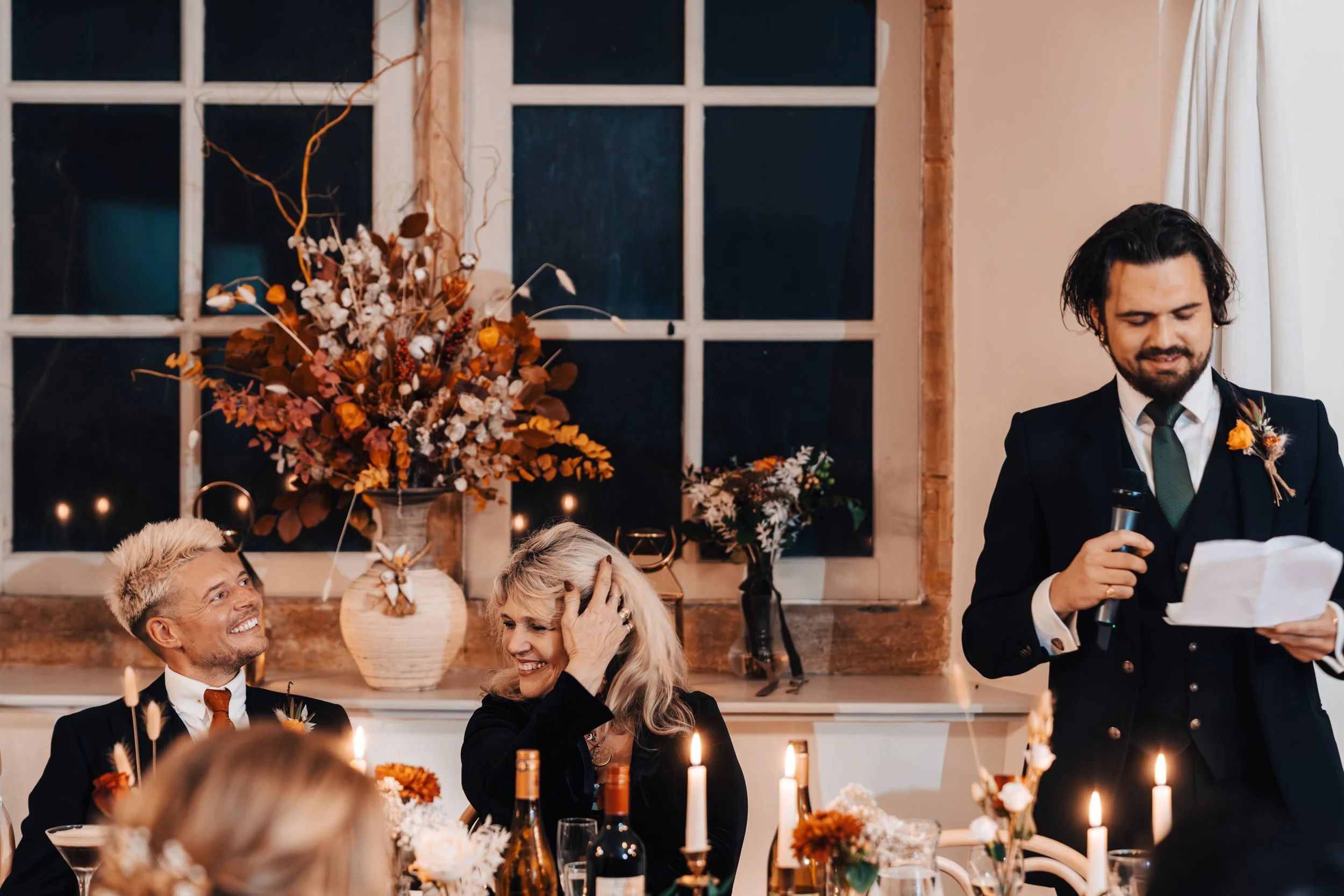 A man giving a speech at a wedding reception, standing near a table with candles and flowers, with two smiling women and a man seated at the table.