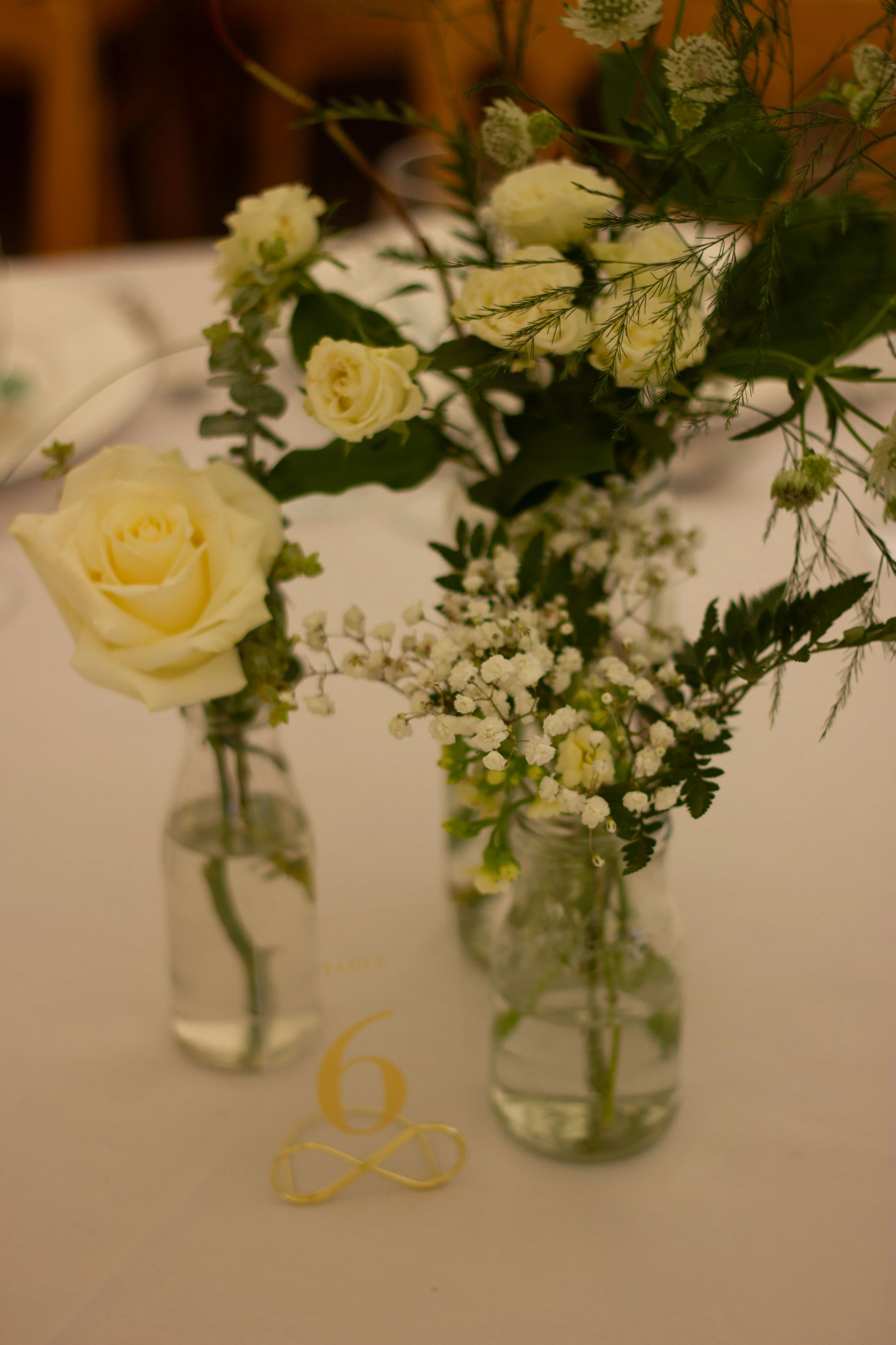 Close-up of two small glass bottles with white roses and greenery, placed on a table with a gold table number 6 and a gold infinity symbol decoration.