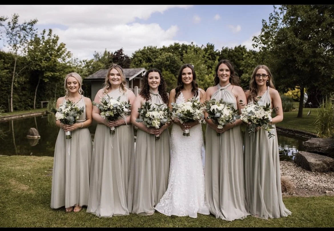 Six women standing outdoors by a pond, dressed in wedding and bridesmaid dresses, holding bouquets, smiling for the camera with trees and a small building in the background.