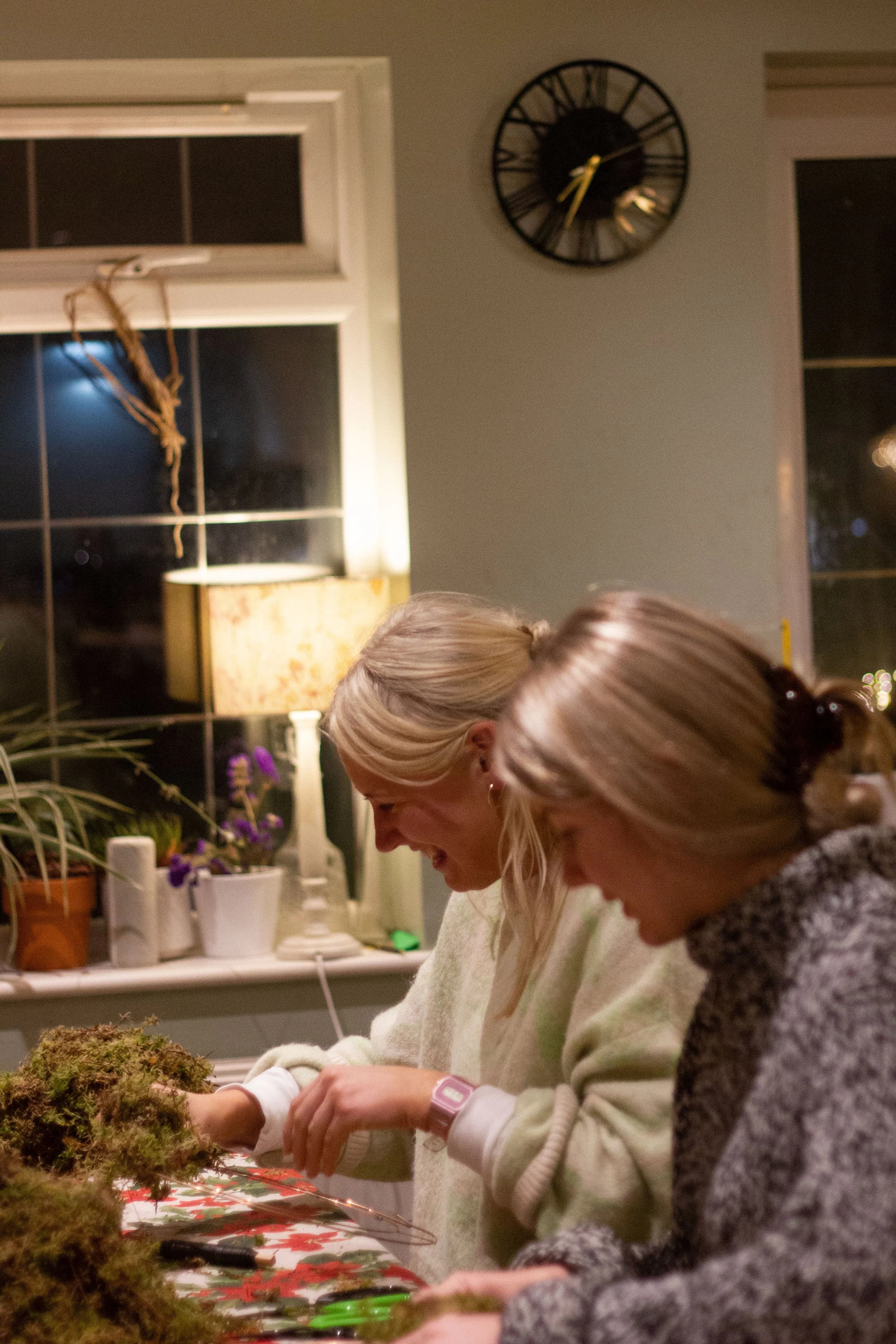 Two women decorating a table with moss and Christmas-themed decor during the evening.