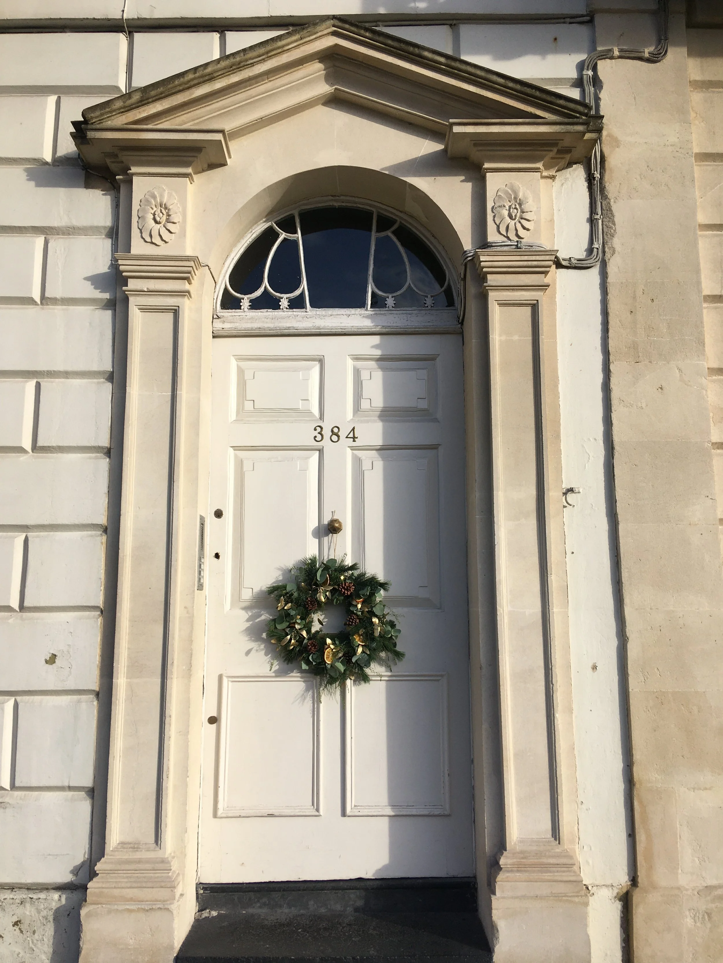 White front door with the number 384, decorated with a green wreath. The door is framed by decorative stone columns and roofwork, with a semi-circular window above.