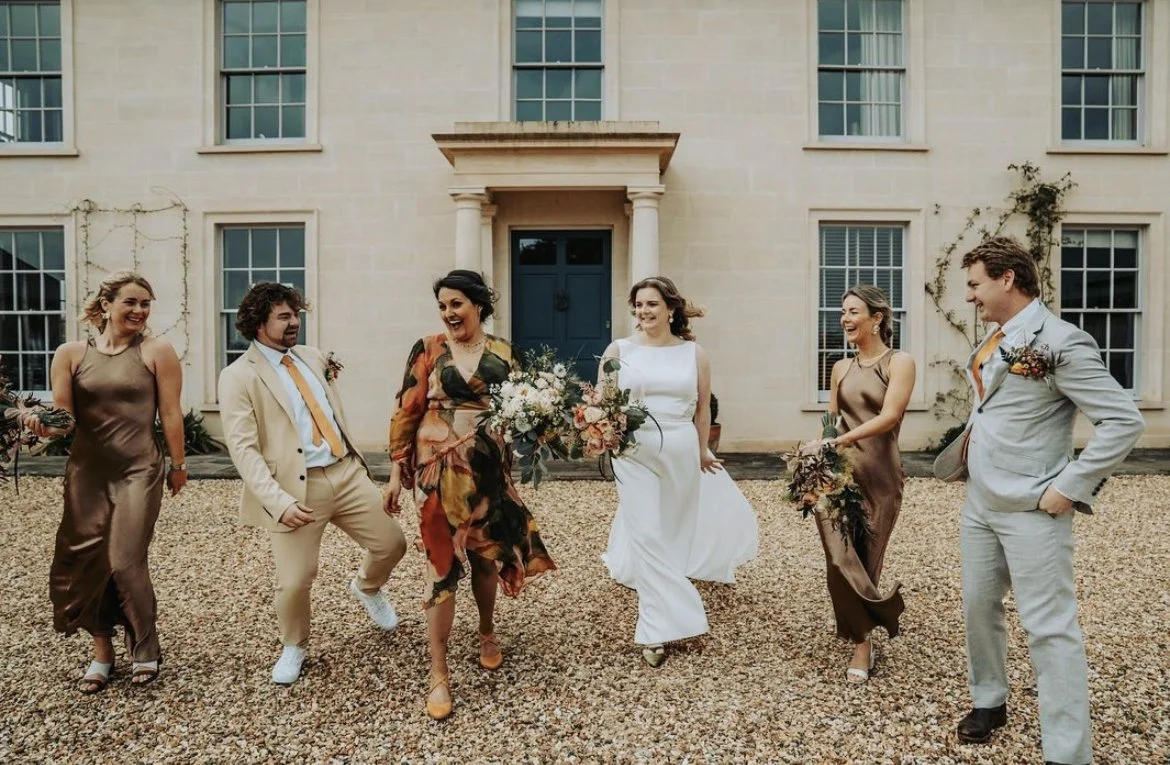 Group of people dressed in formal attire, walking and smiling outside a large building with multiple windows, during a wedding celebration.