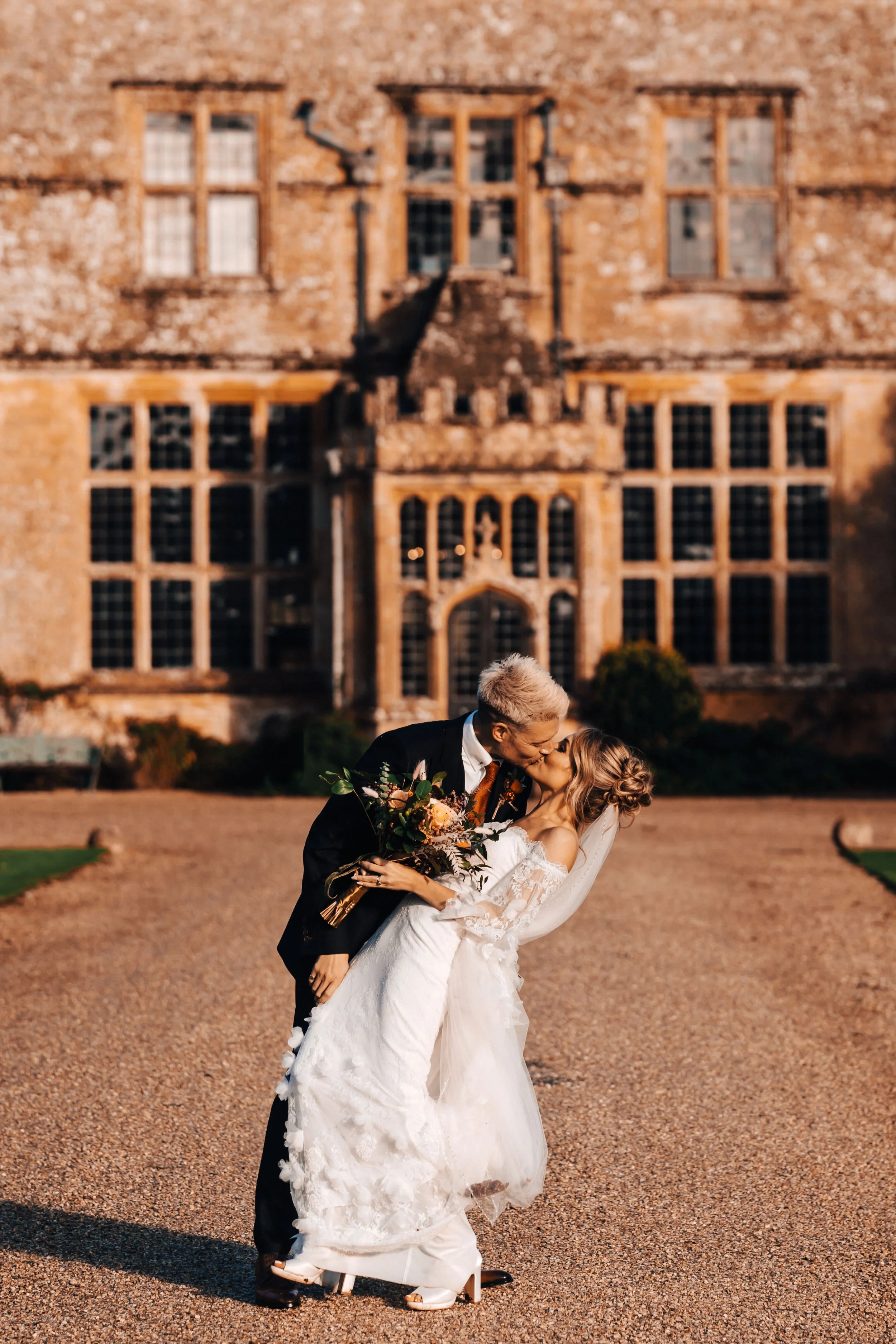 A newlywed couple sharing a kiss during their wedding photoshoot outside a historic brick mansion. The groom is dressed in a black tuxedo and the bride in a white lace wedding gown, holding a bouquet of flowers.